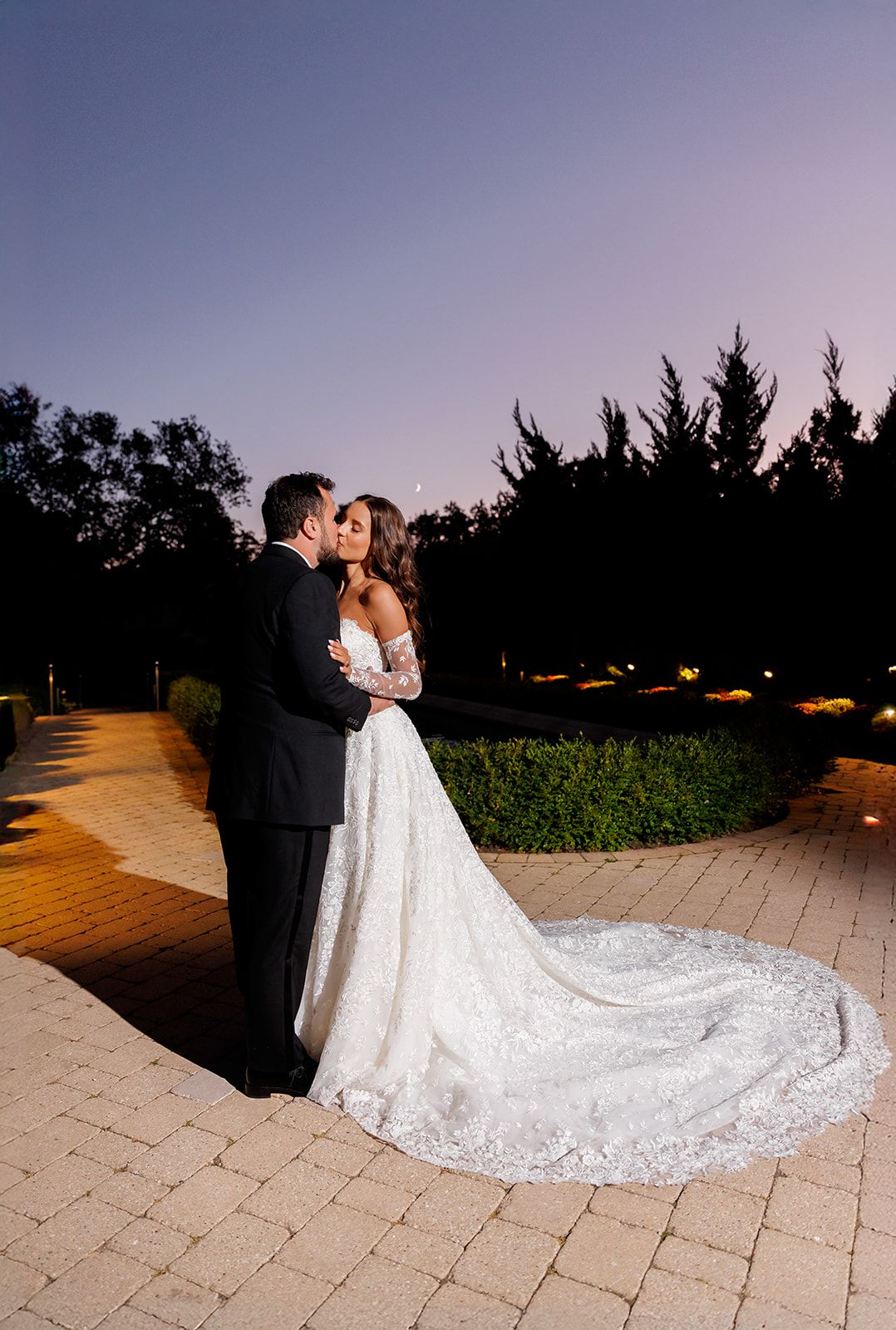 Bride and groom share a romantic kiss at dusk, her lace gown train spread across a stone patio.