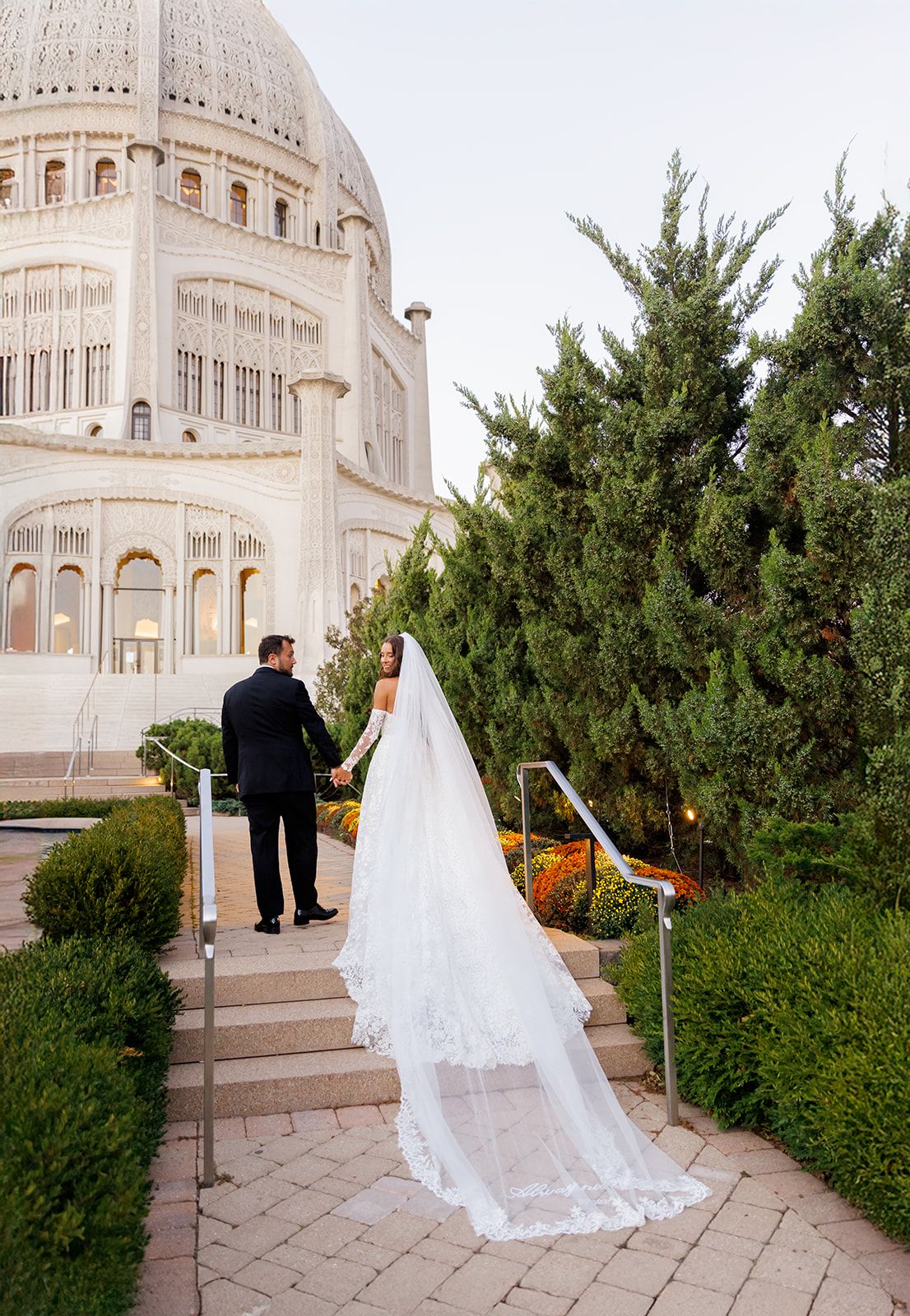 Bride and groom hold hands on garden steps before ornate domed Bahai Temple, bride wearing cathedral lace veil.