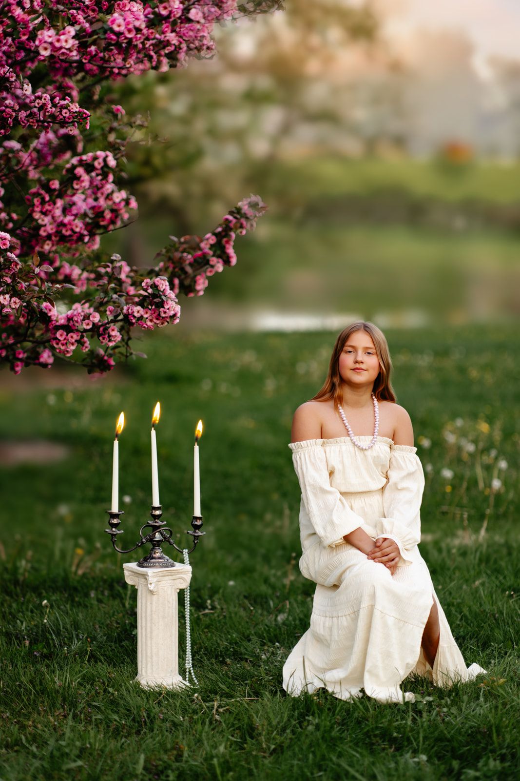 Young girl in white off-shoulder dress sits on grass near lit candles and pink blooming tree at sunset.