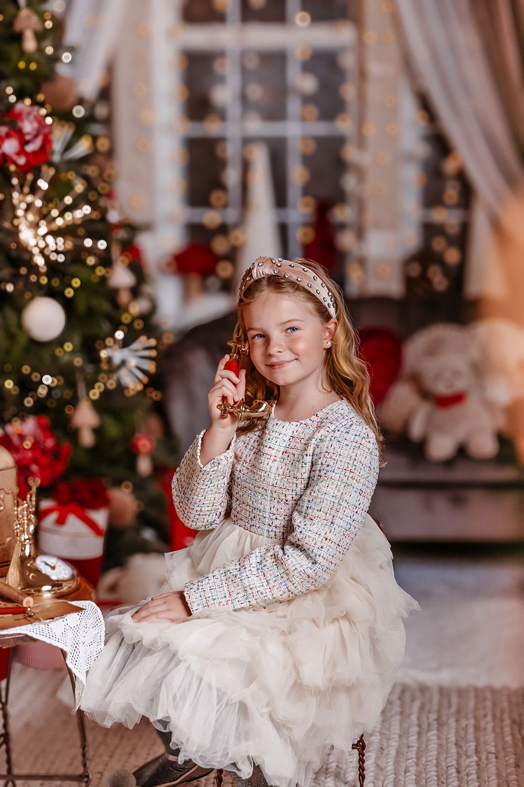 Young girl in elegant dress smiling by decorated Christmas tree with festive lights and gifts.