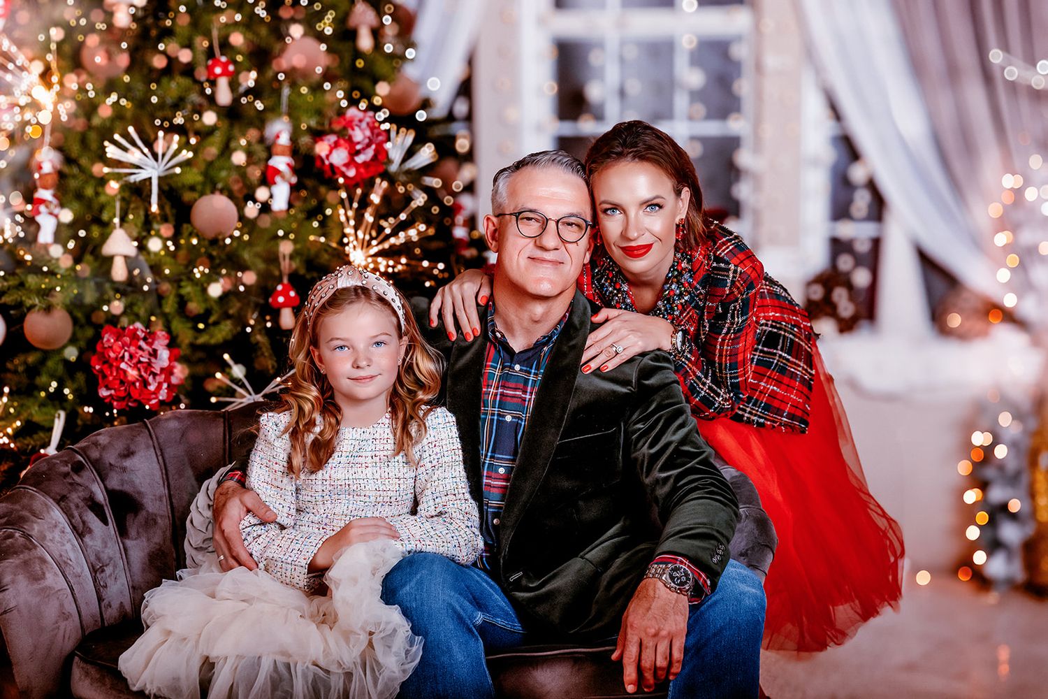 Family of three posing by decorated Christmas tree, grandfather, grandmother in red, and granddaughter in white dress.