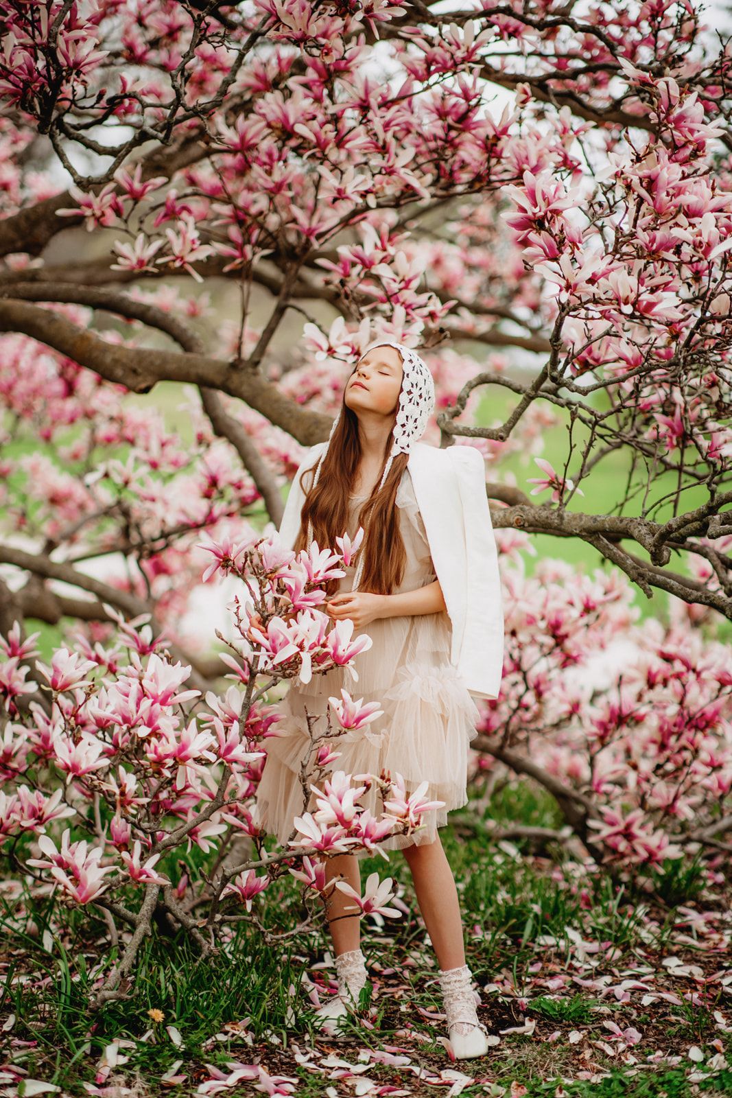 Young girl in white dress and bonnet standing among pink magnolia blossoms in spring garden.