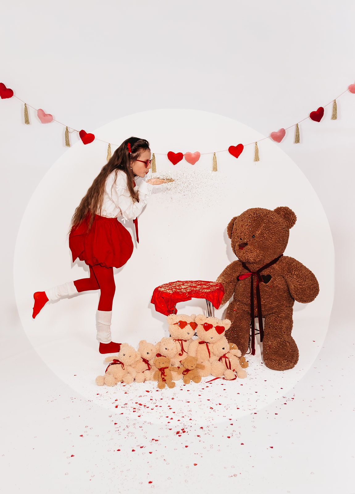 Girl blowing glitter near teddy bears and a large brown bear, Valentine's Day themed photo with heart garland.