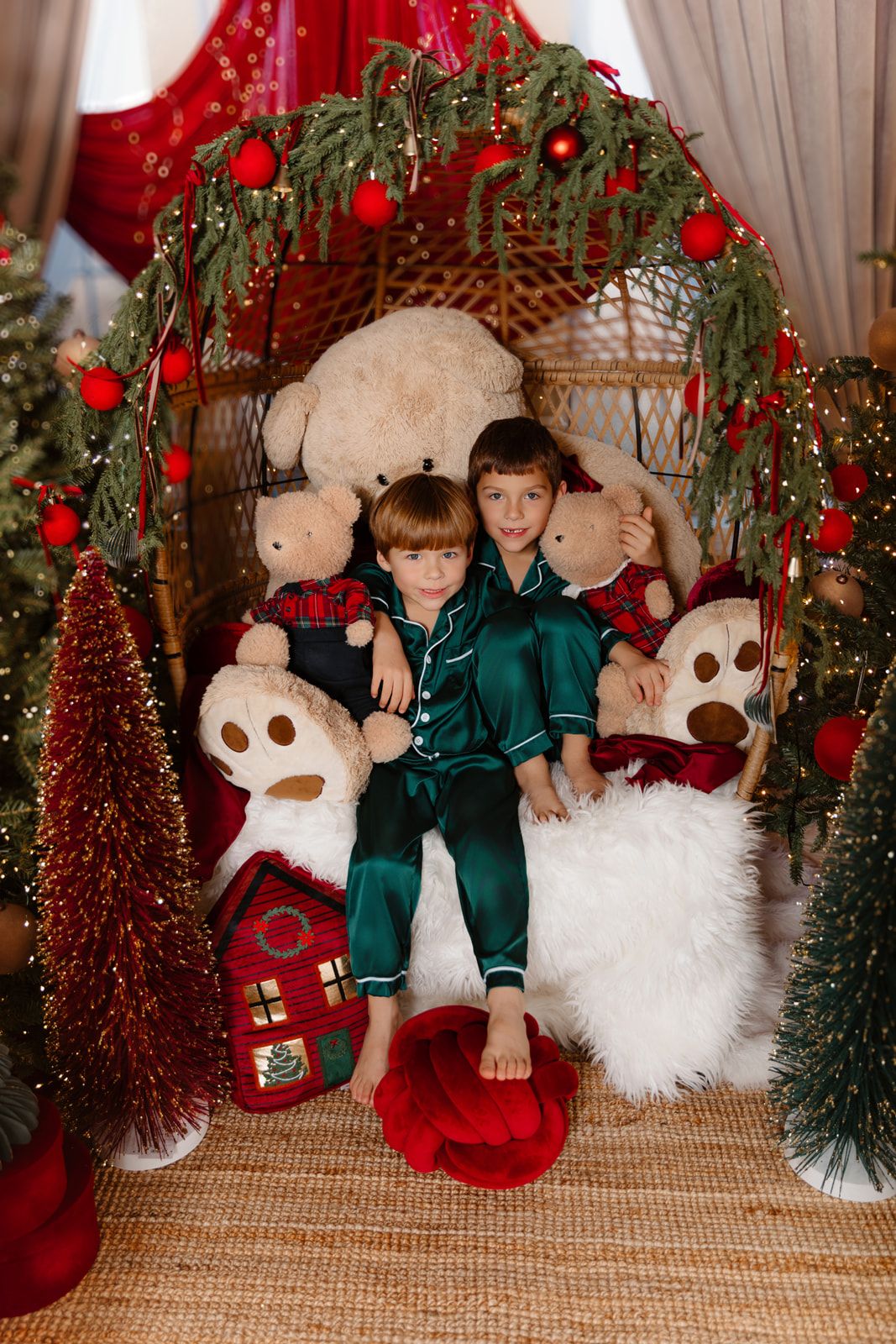 Two children in green pajamas sit in a decorated wicker chair surrounded by teddy bears and Christmas trees.