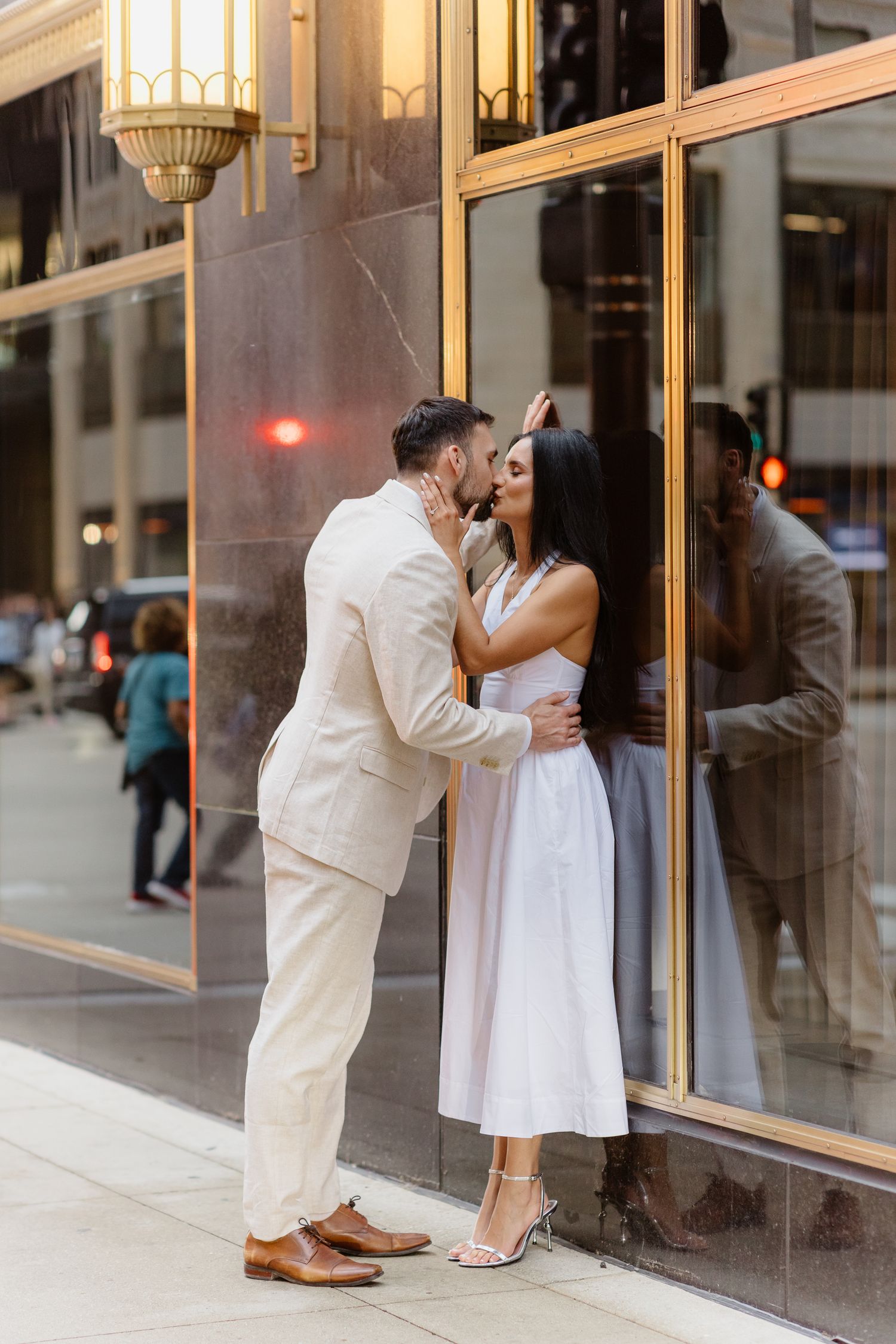 Man in beige suit kissing woman in white dress against luxury building exterior on city street.