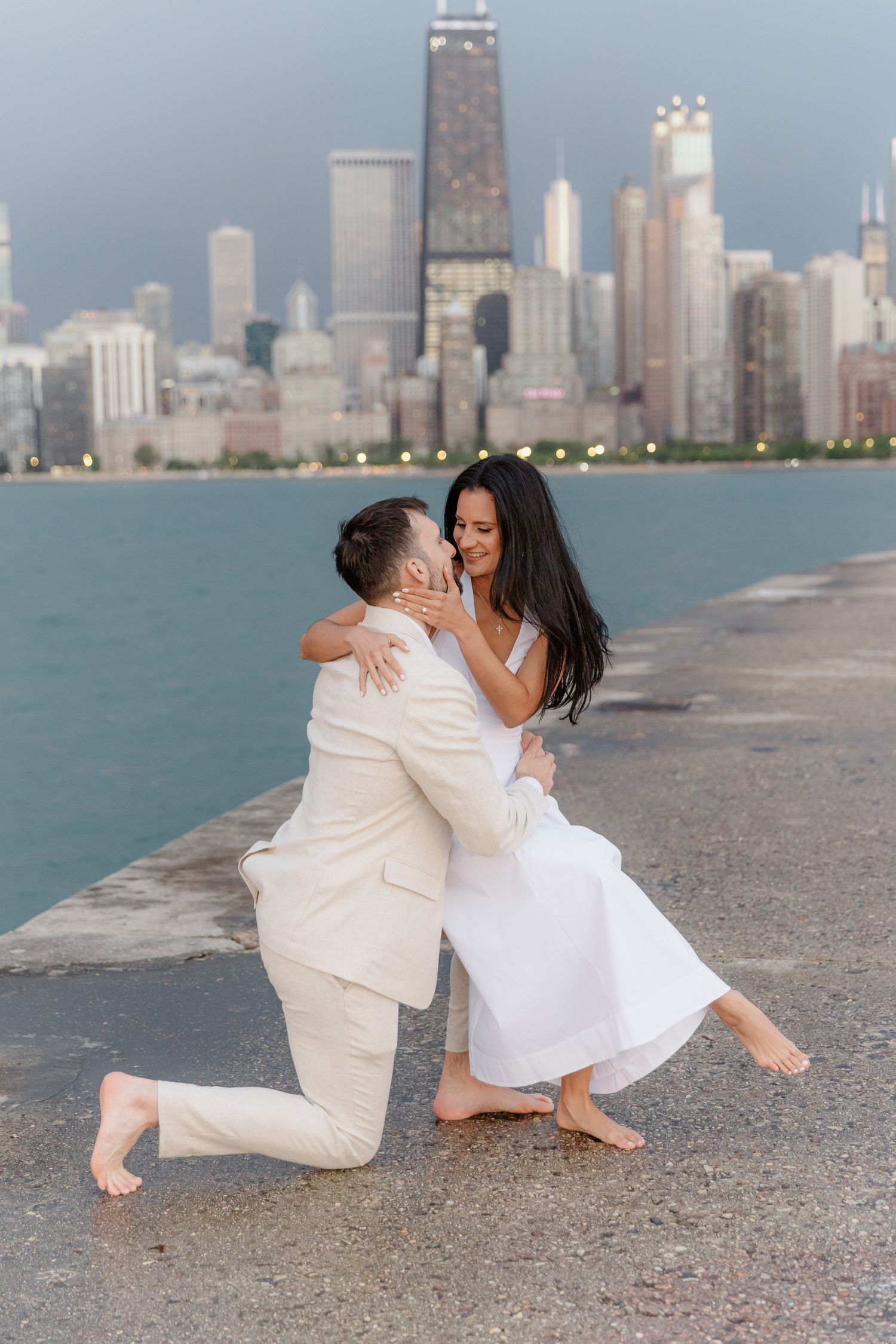 Man on one knee proposing to woman on Chicago lakefront pier with city skyline backdrop.