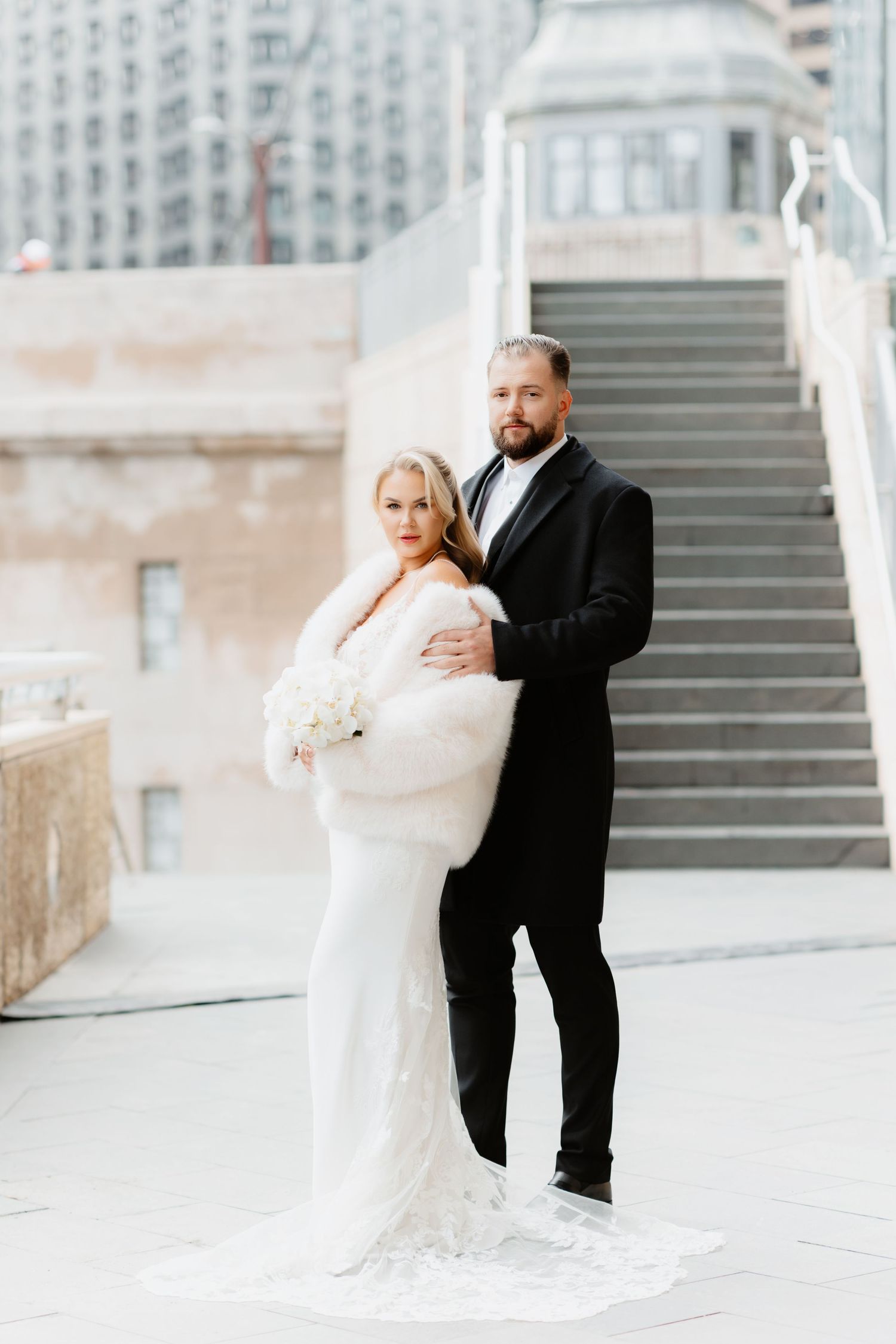 Bride in white fur stole and groom in black tuxedo pose on urban steps in Chicago.