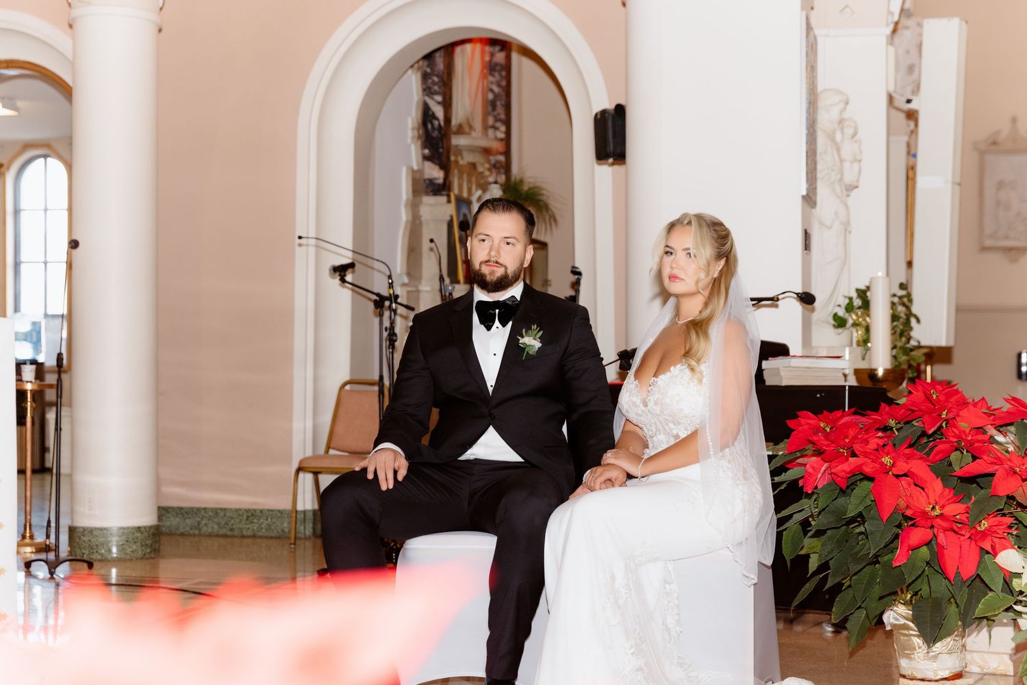 Groom in black tuxedo and bride in white gown seated at church altar with poinsettias.