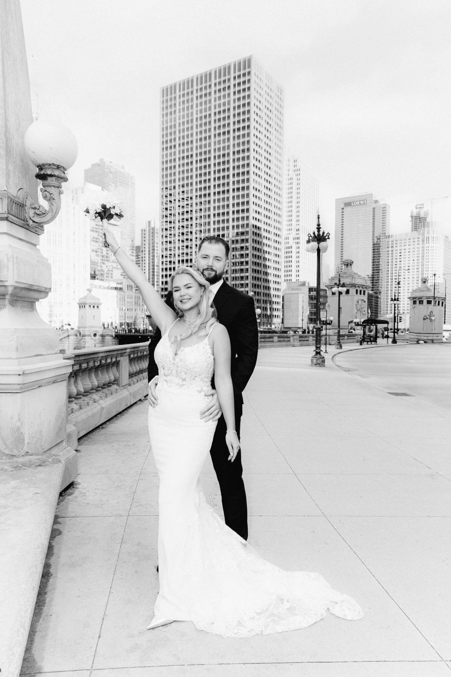 Black and white photo of bride raising bouquet with groom on a Chicago bridge with city skyline.