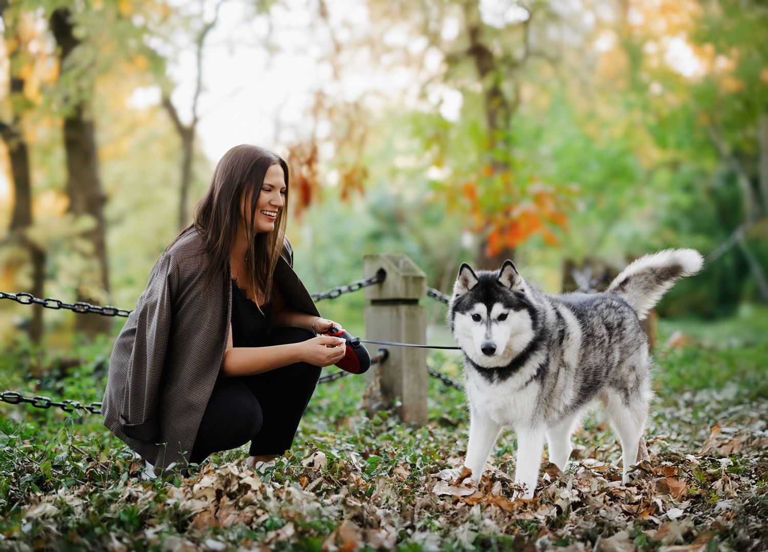 Woman crouching beside a Siberian Husky dog on a leash in an autumn forest setting.