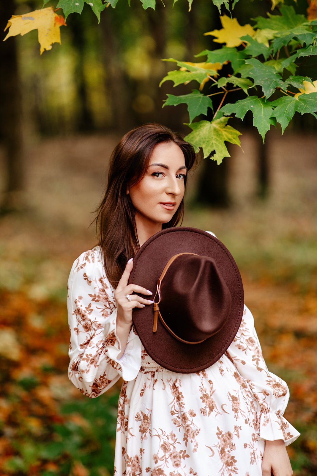 Woman in floral dress holding brown wide-brim hat in autumn forest with colorful maple leaves.