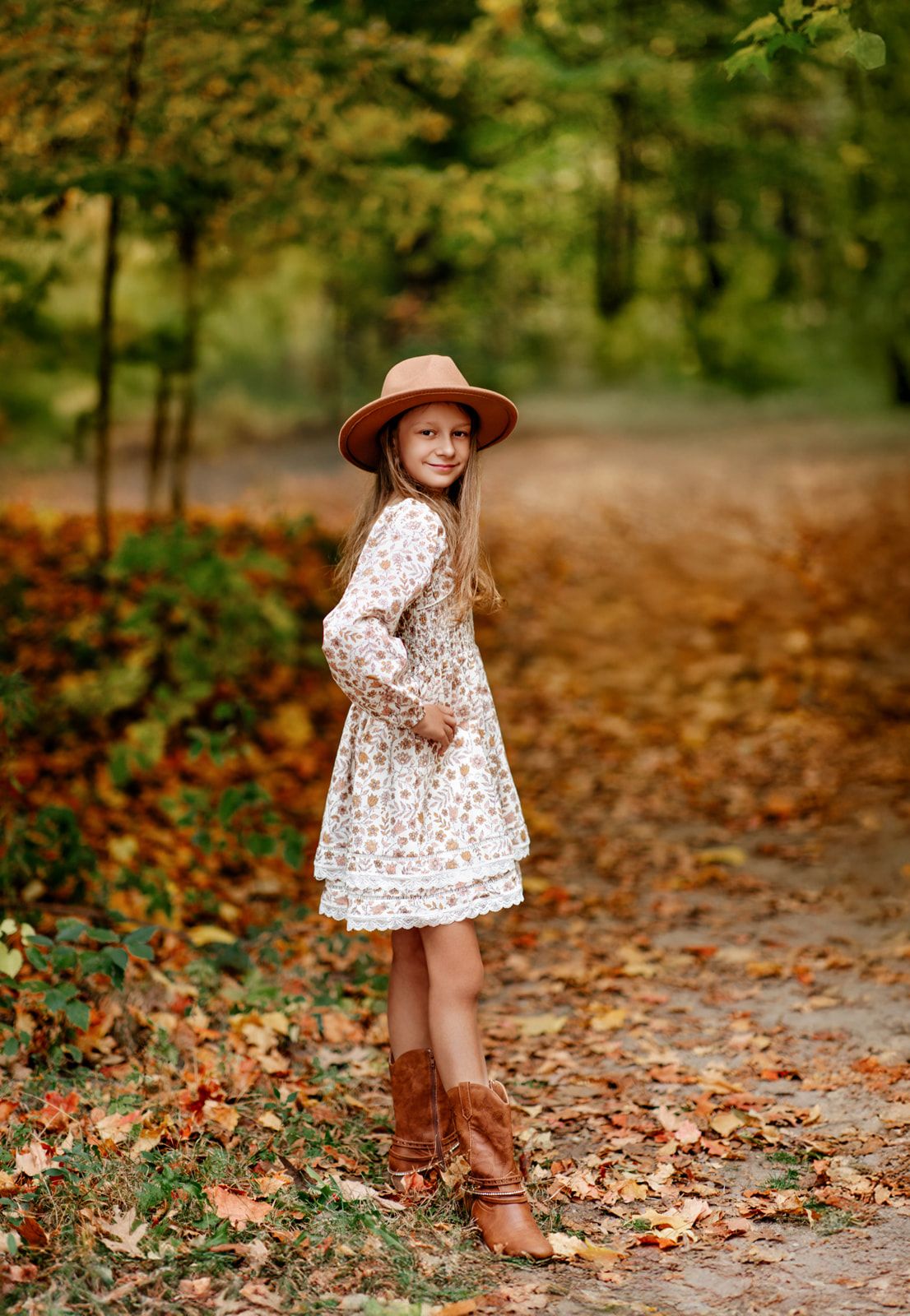 Young girl in white floral dress, tan wide-brim hat and brown cowboy boots in autumn forest path.