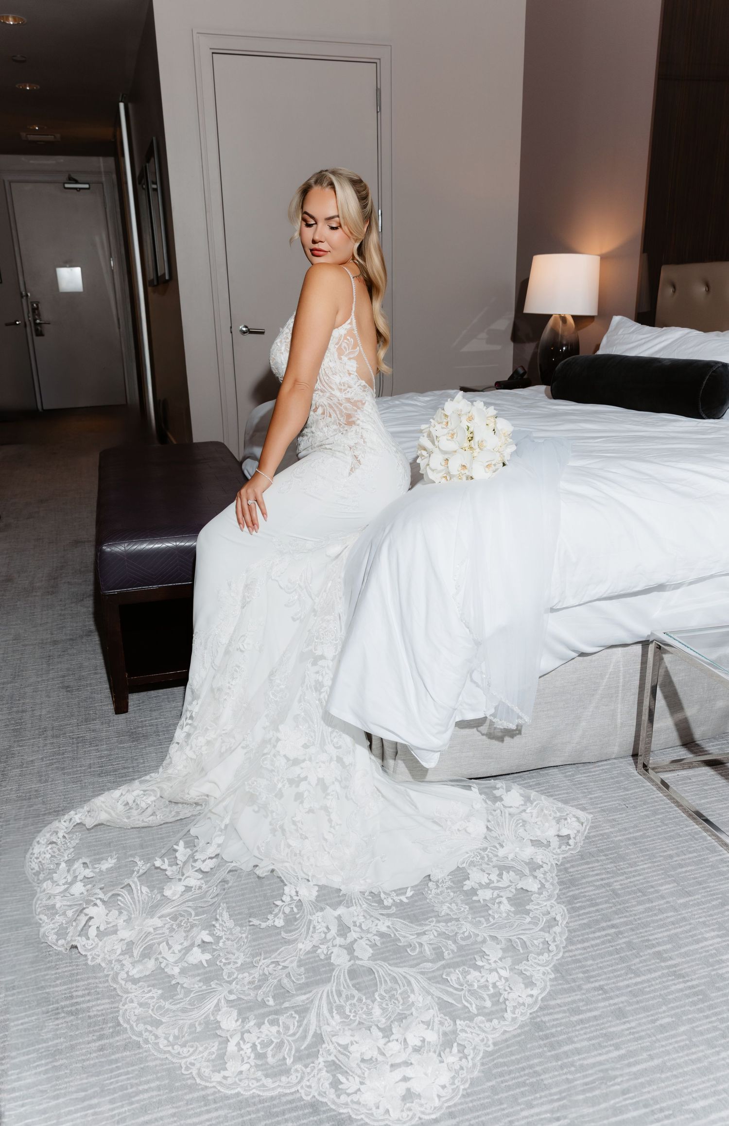 Beautiful blonde bride in sleeveless lace mermaid gown with floral train holds white bouquet on hotel bed.