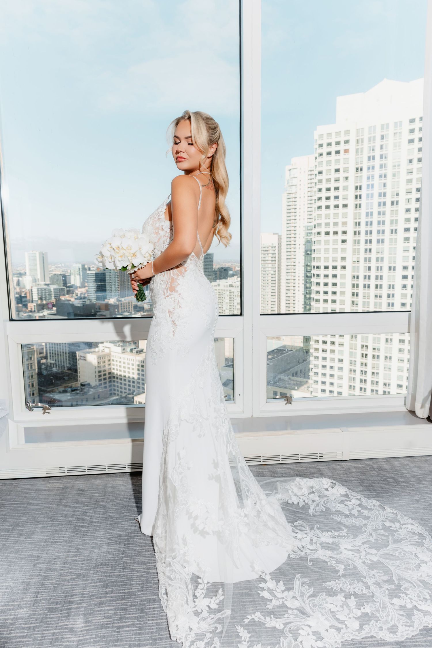 Elegant bride holding white bouquet in fitted lace gown with cathedral train before urban skyline windows.