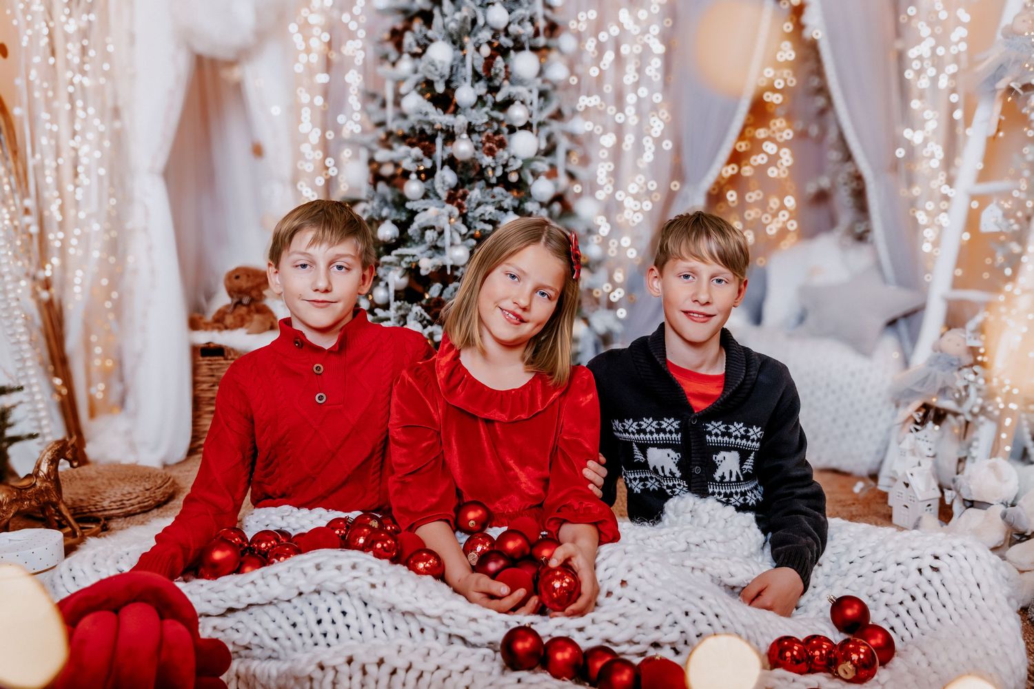 Three children in red Christmas outfits sit together holding ornaments near a glowing holiday tree.