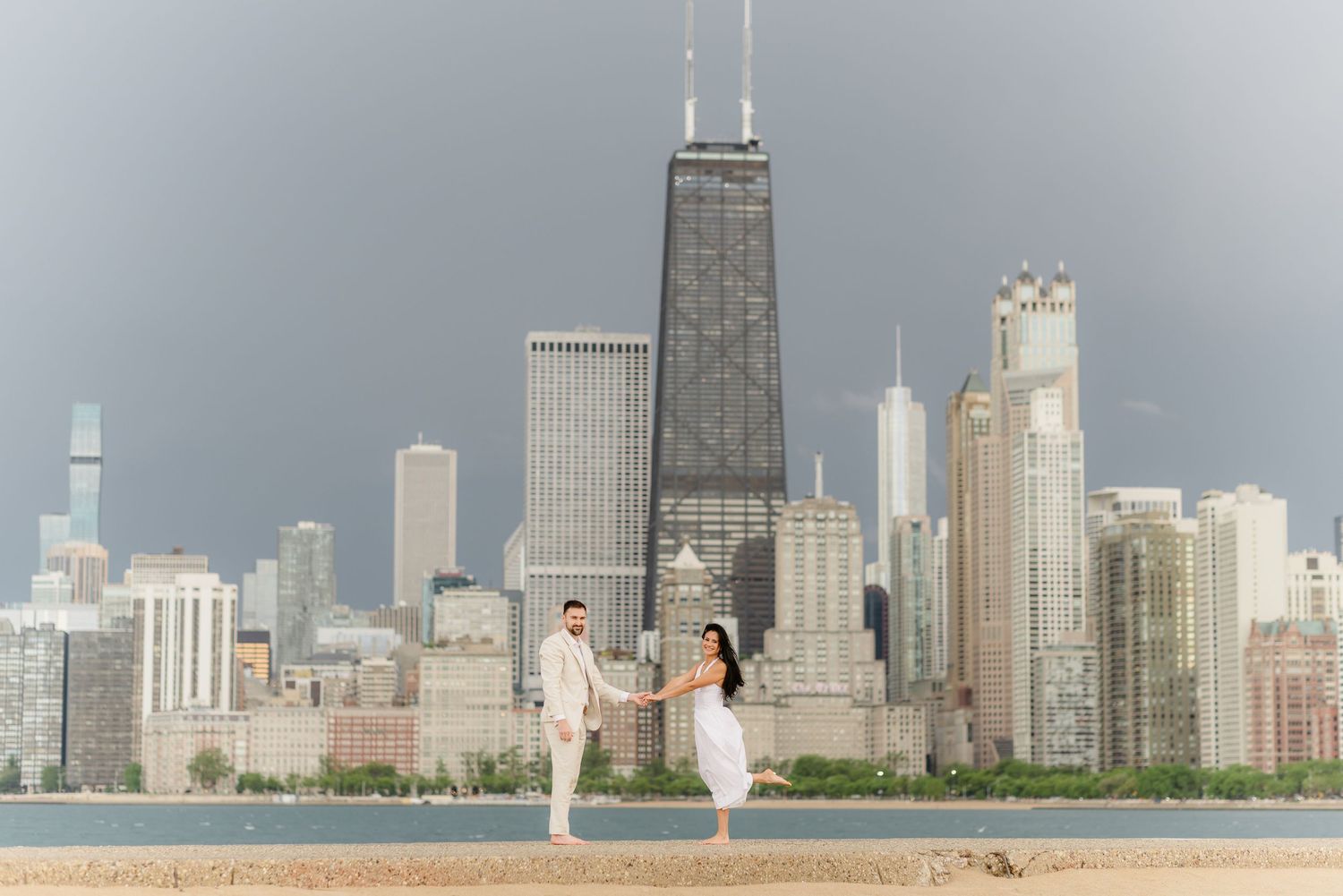 Barefoot couple holding hands walking along Chicago lakefront with John Hancock Tower skyline view.