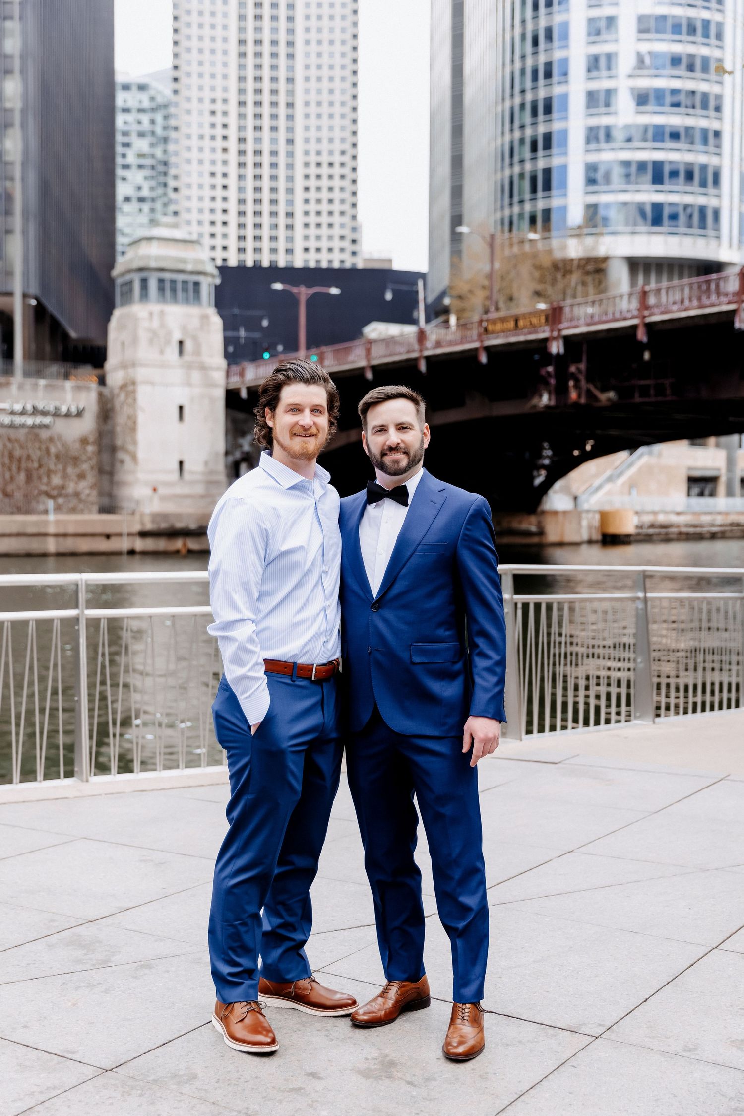 Two men in navy blue suits pose together on an urban waterfront with a city bridge behind them.