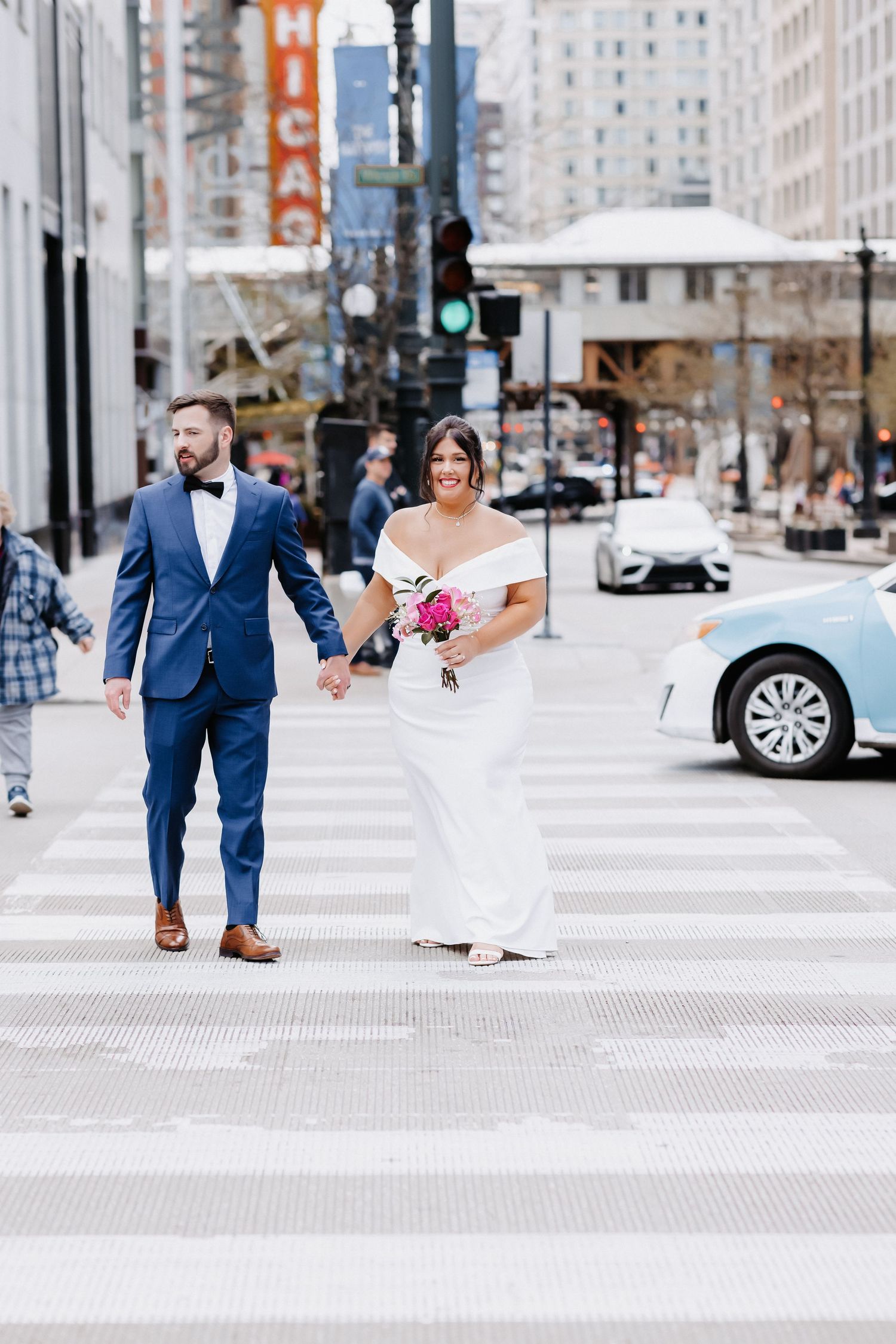Wedding couple holds hands crossing Chicago crosswalk, bride in white gown with pink bouquet, groom in navy suit.