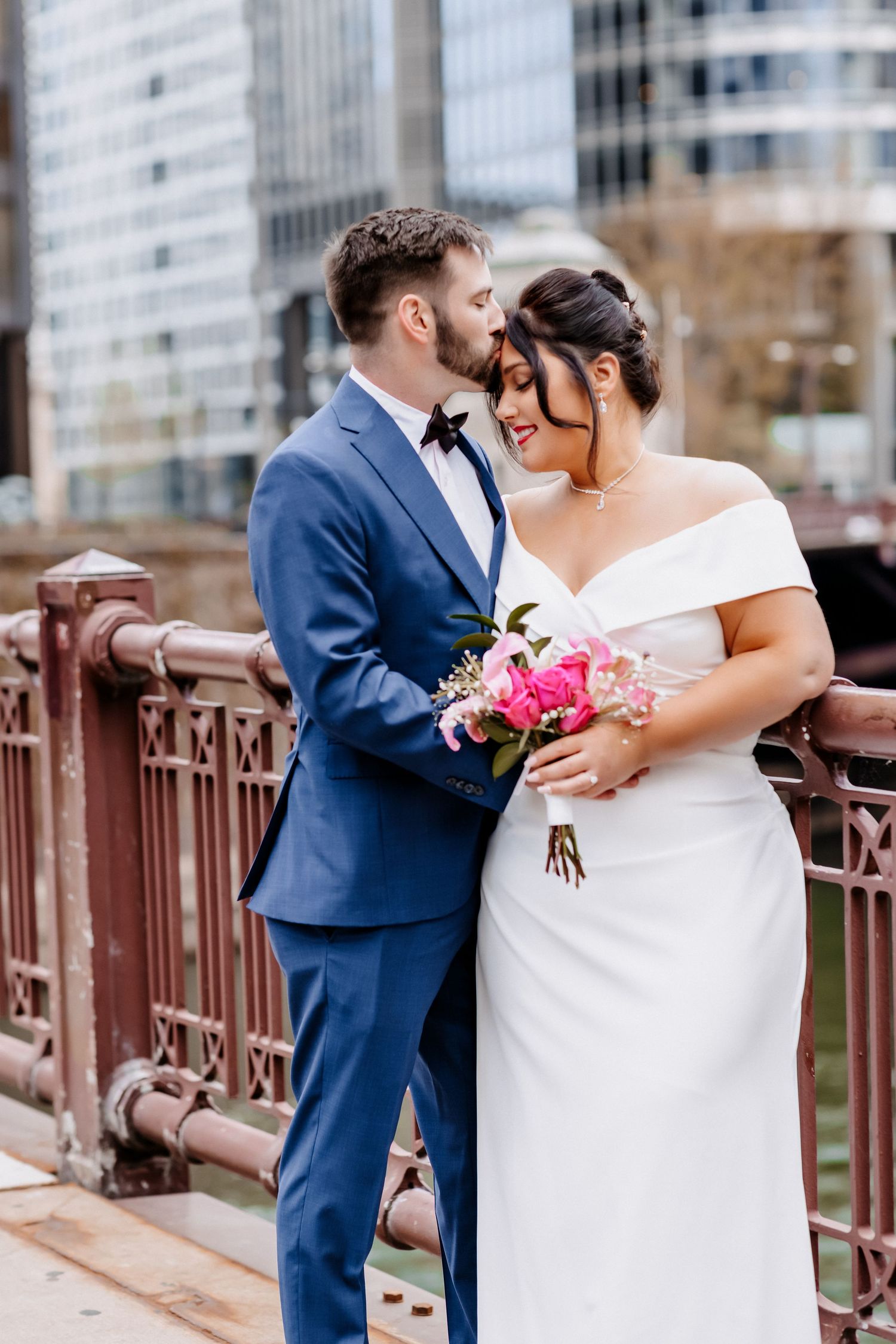 Bride and groom share a romantic moment on a bridge, she holds pink bouquet, he wears blue suit, city skyline behind.