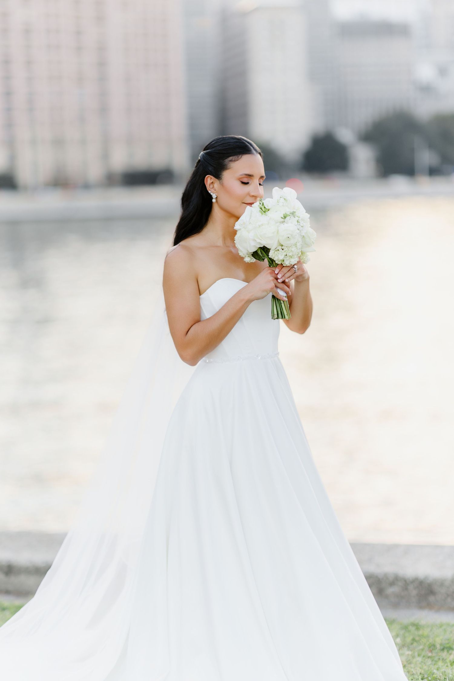 Bride in strapless white gown holds white floral bouquet near waterfront with city skyline backdrop.