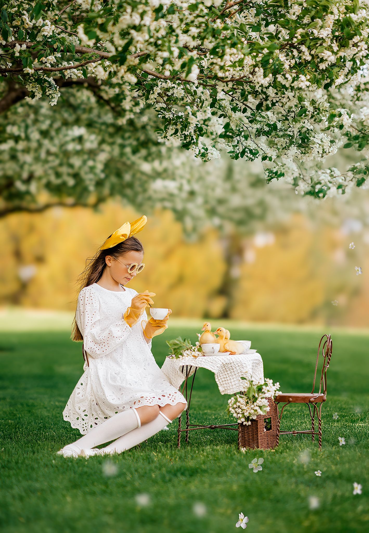 Girl in white dress and yellow bow enjoys tea party under blooming apple tree on lush green lawn.