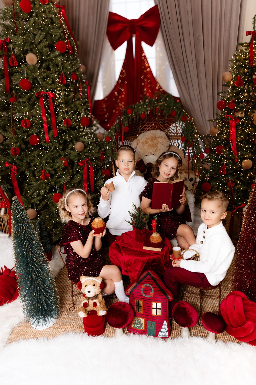 Four children in red and white outfits sit among decorated Christmas trees with ornaments, gifts, and holiday decorations.