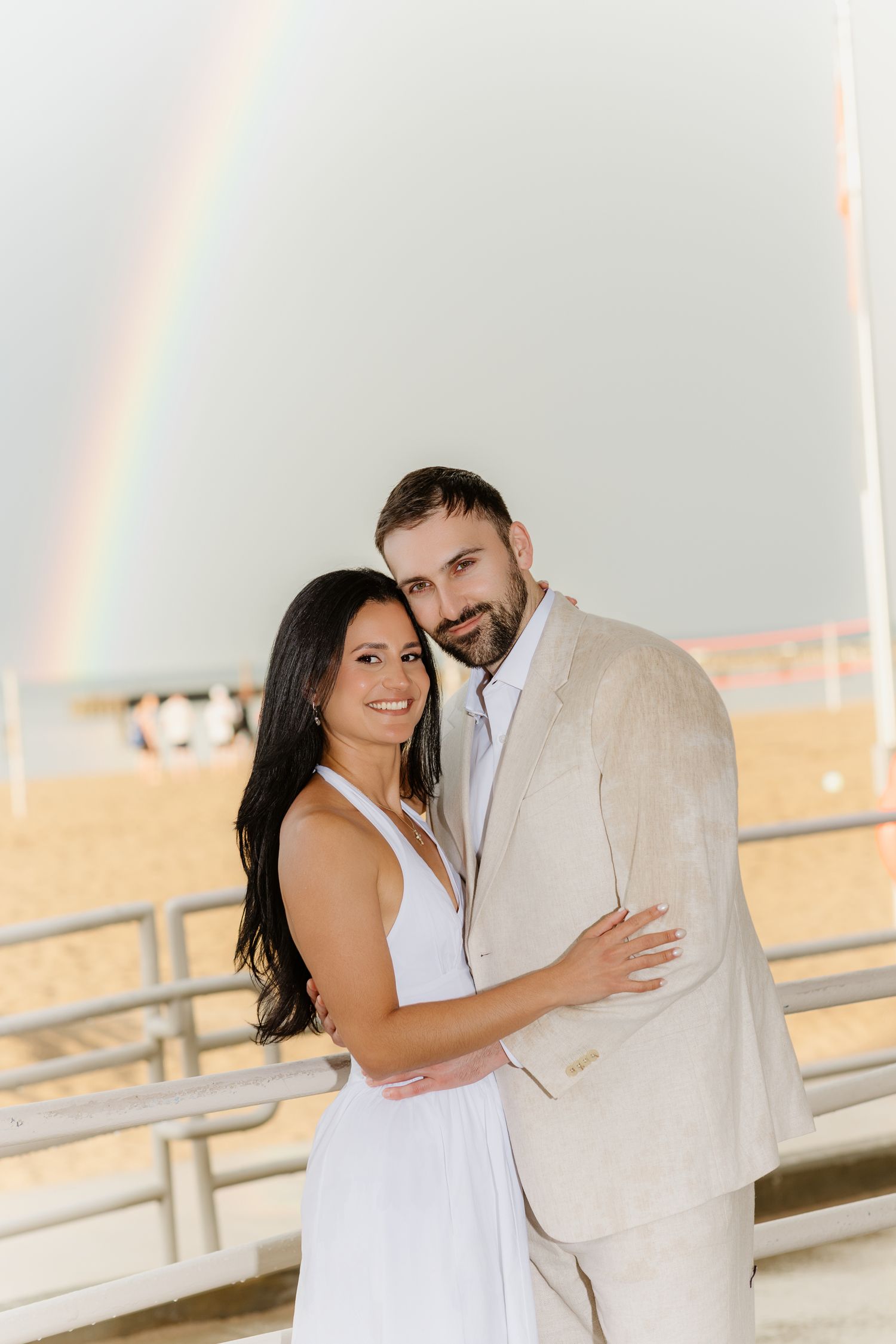 Happy engaged couple in white dress and tan suit posing with a rainbow over the beach behind them.