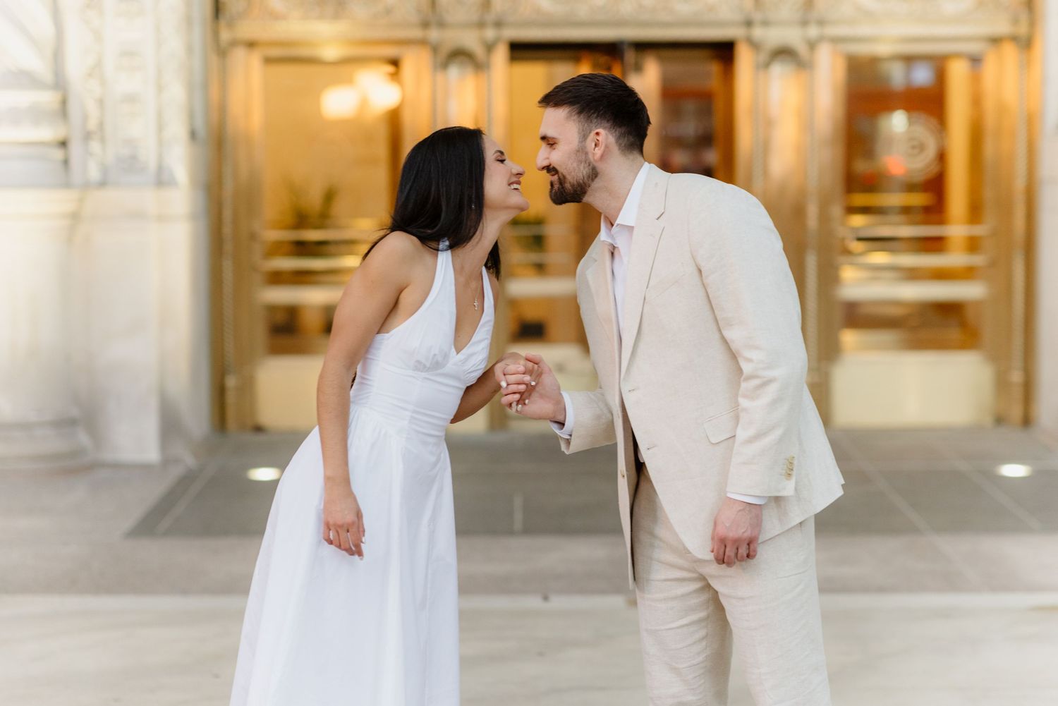 Smiling couple in white dress and beige suit holding hands outside golden building.