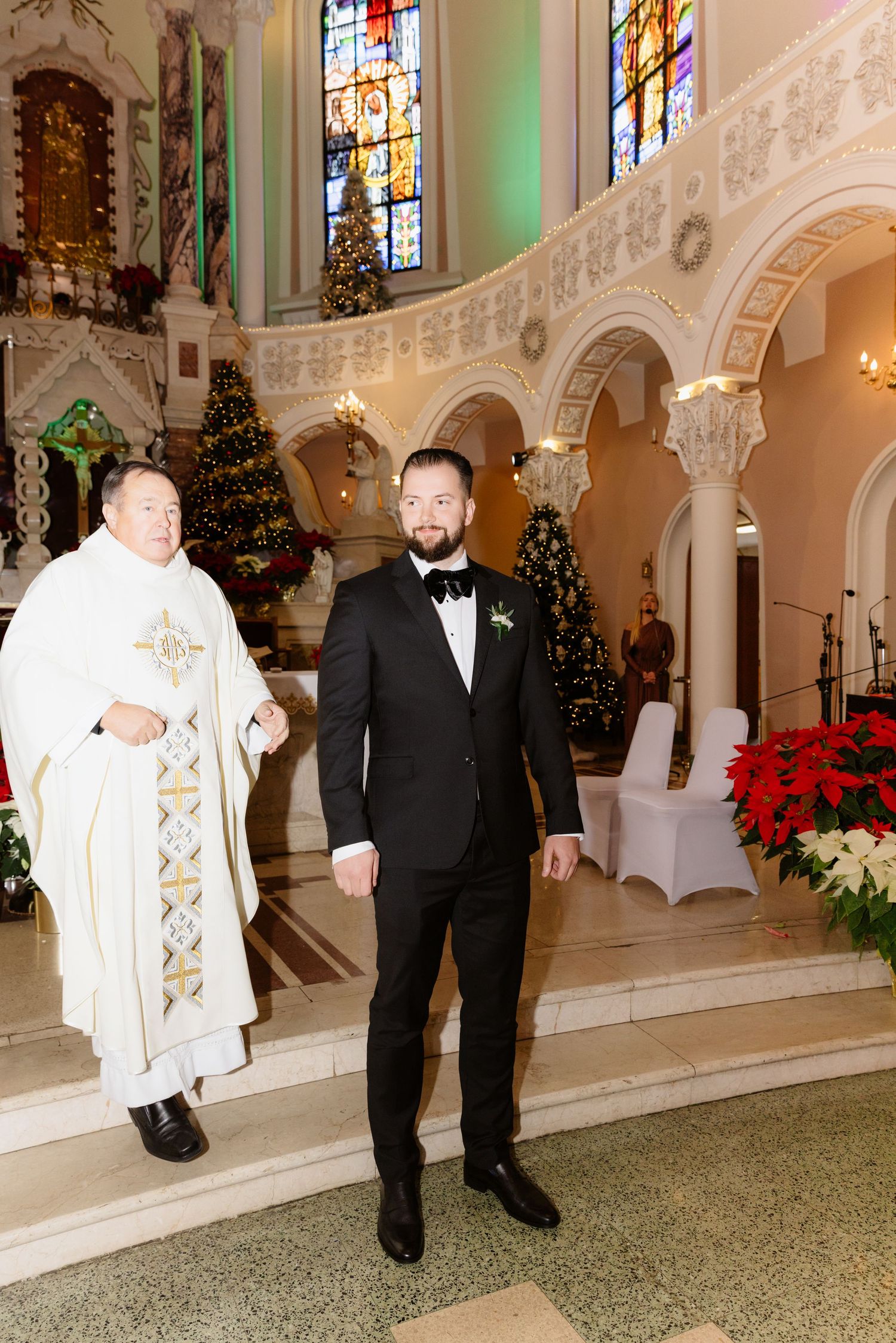 A bearded groom in a black tuxedo stands at the church altar beside a priest in white vestments.