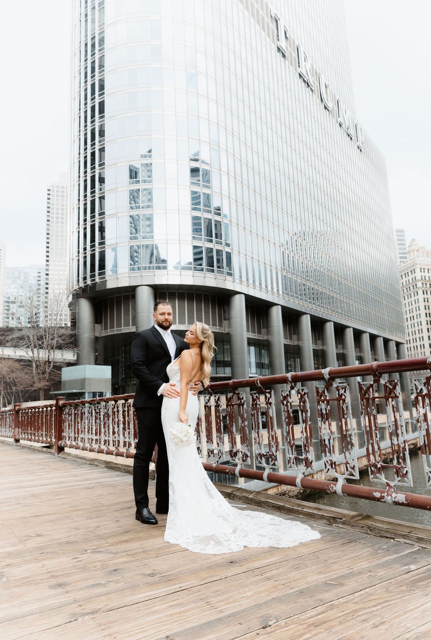 Bride in backless lace gown and groom embrace on Chicago Riverwalk with skyscrapers.