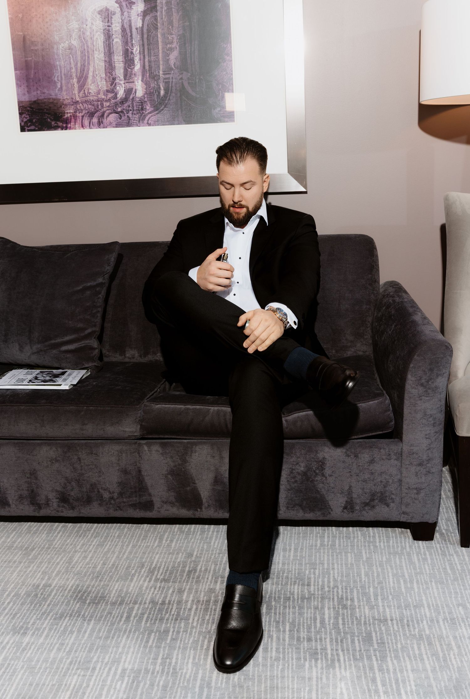 Groom in black suit adjusts cufflinks while seated on gray sofa during wedding day preparations.