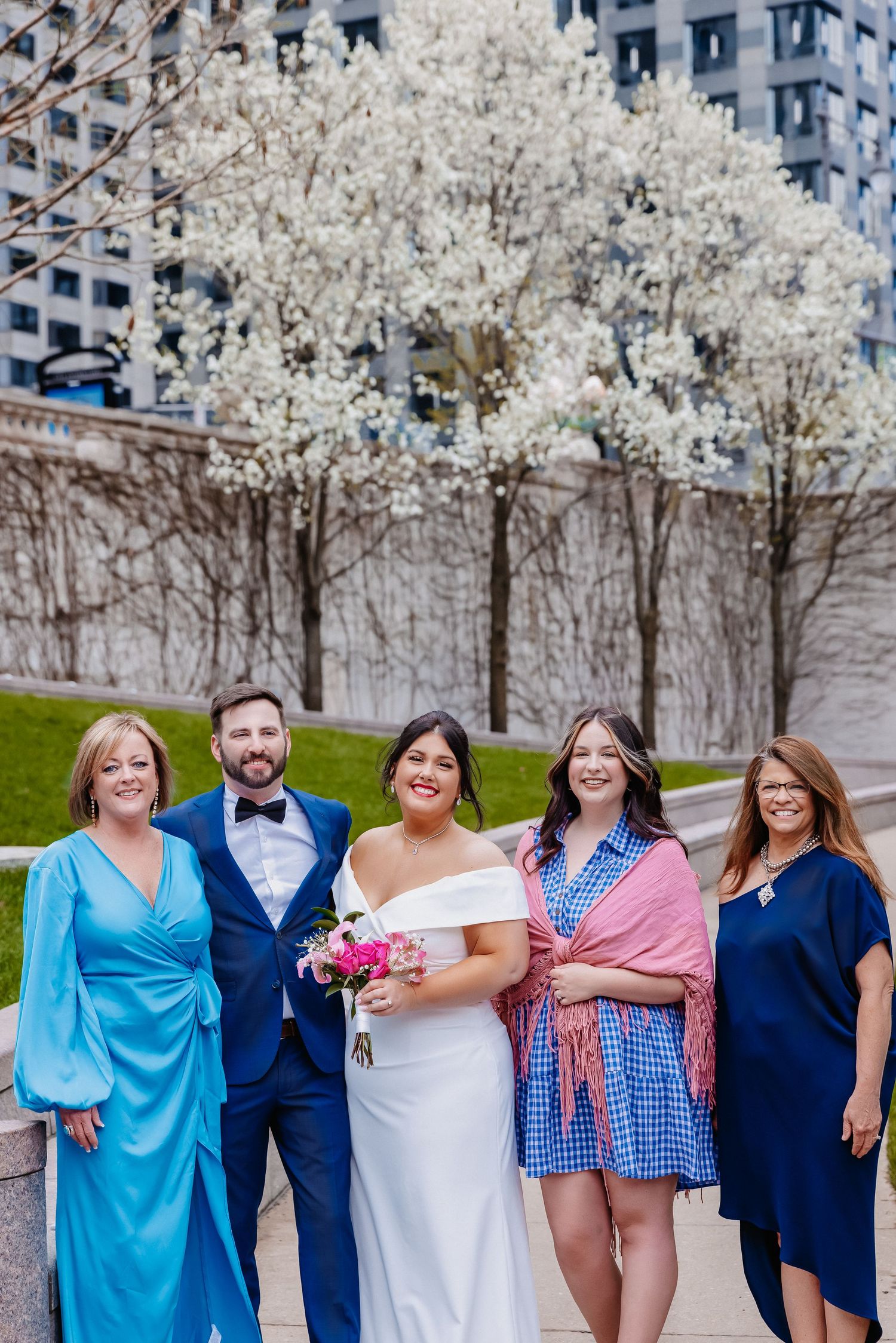 Wedding group of five poses outdoors near blooming trees, bride holding vibrant pink bouquet.