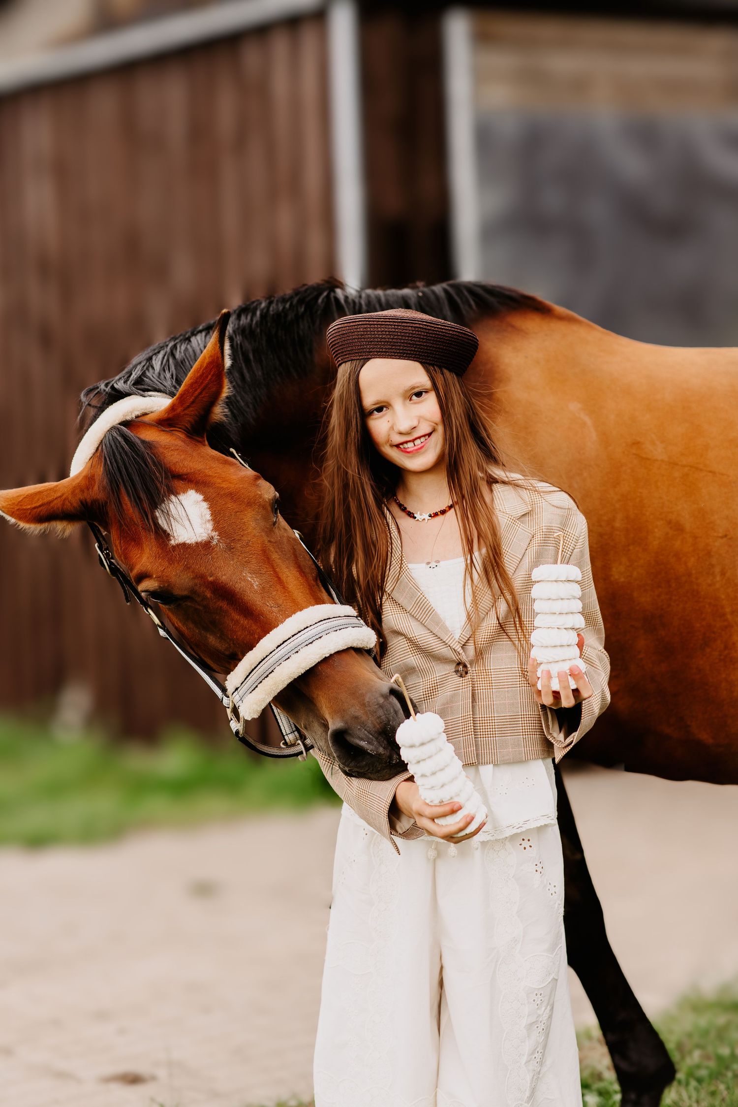 Happy young girl in brown hat and beige jacket standing beside a chestnut horse while holding stacked white treats.