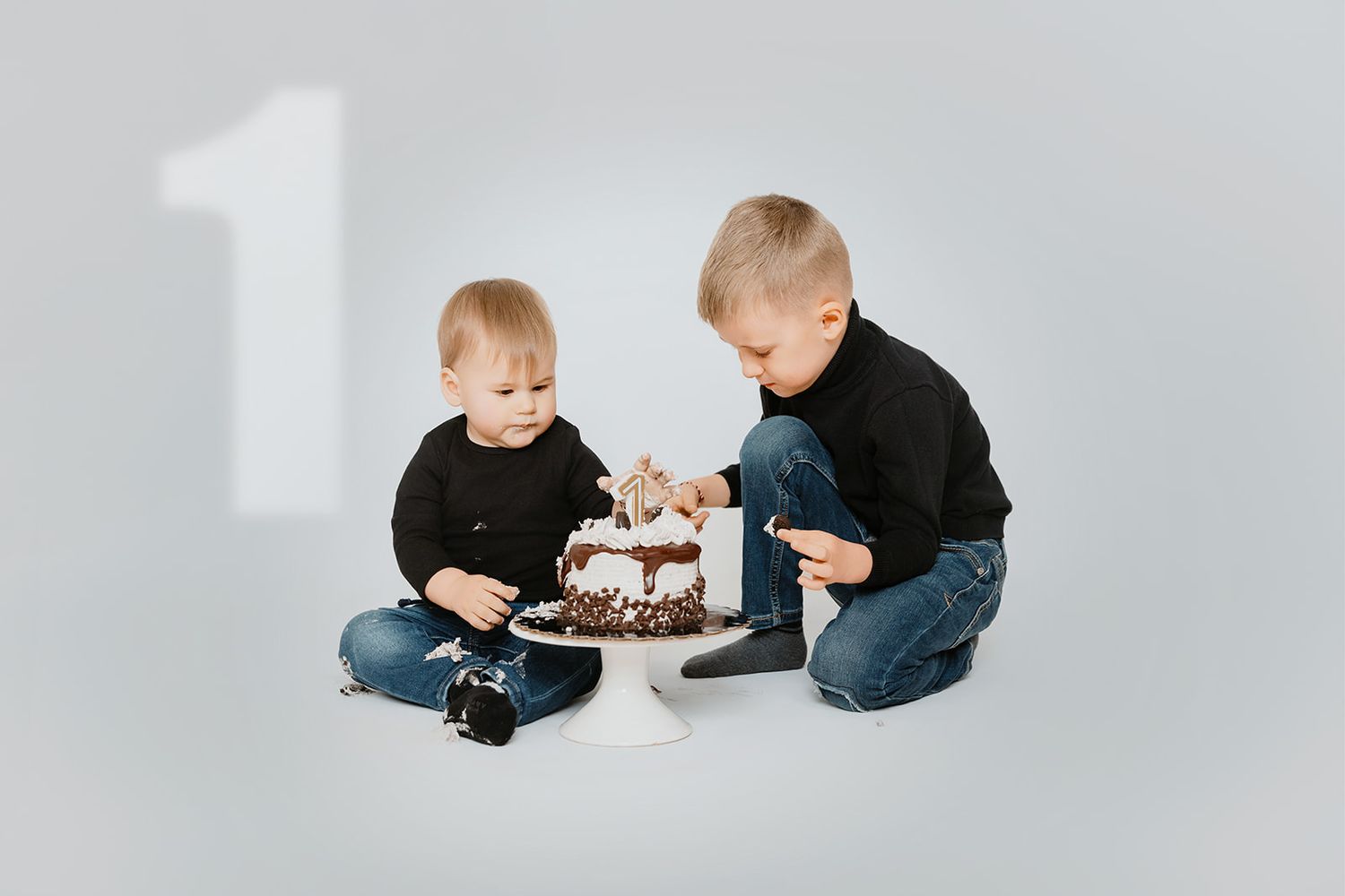 Two young brothers in matching black outfits examine a decorated birthday cake on white floor.