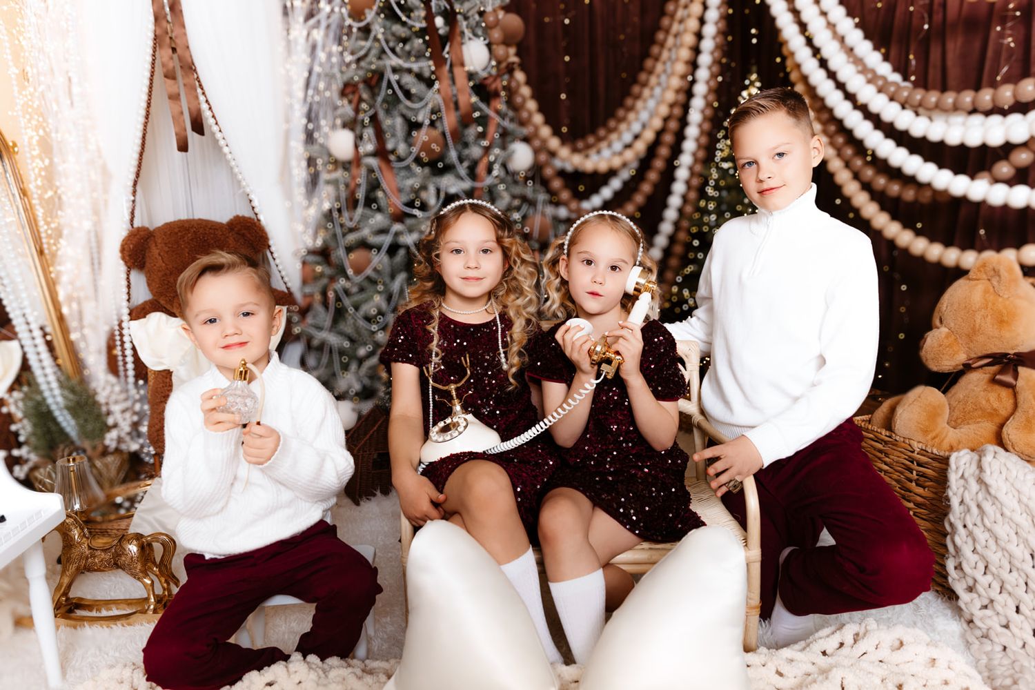 Four children pose together in festive holiday outfits near a decorated Christmas tree with garlands and ornaments.