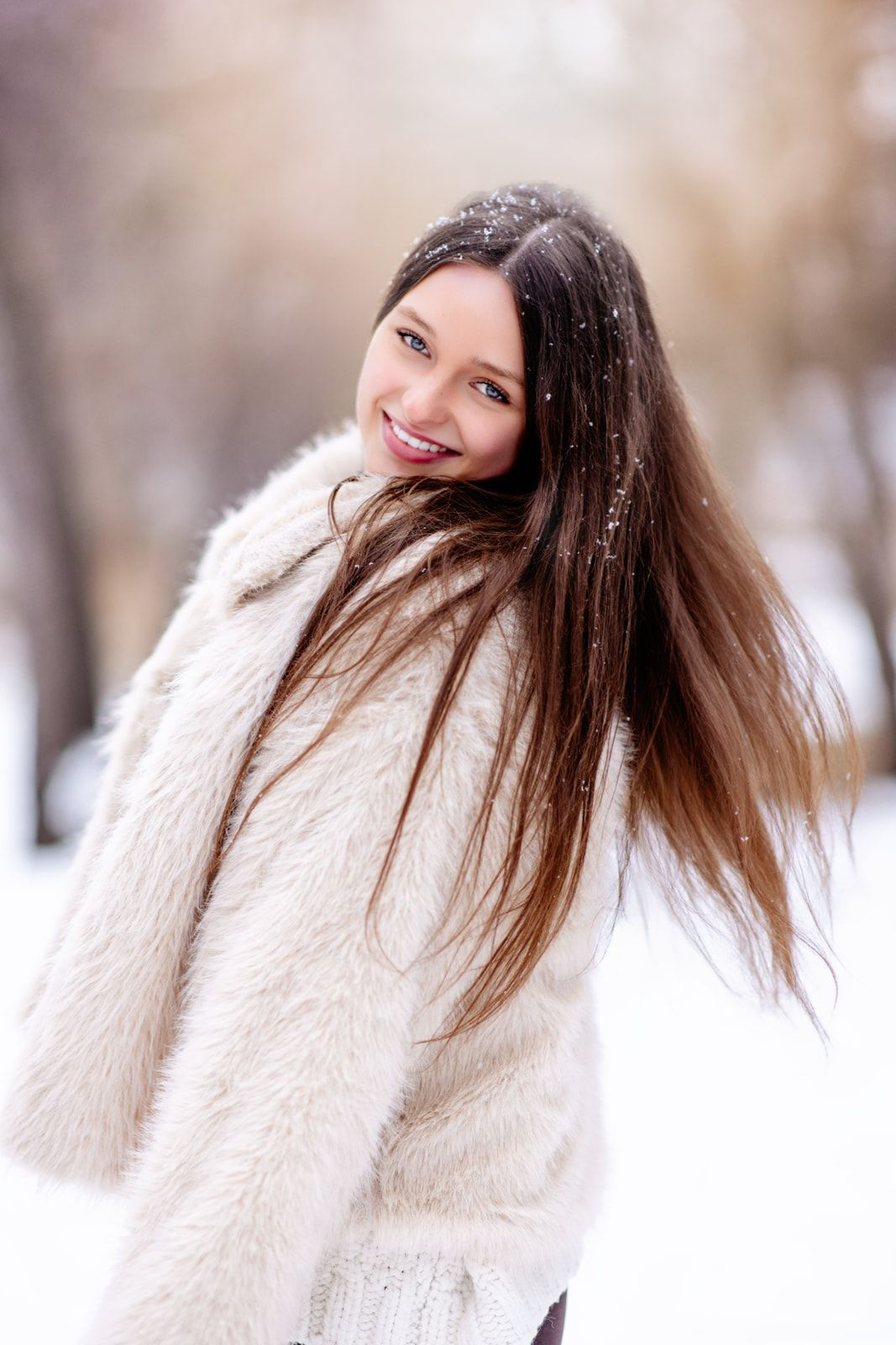 Joyful young woman in cream fur coat with flowing hair posing in snowy winter park.