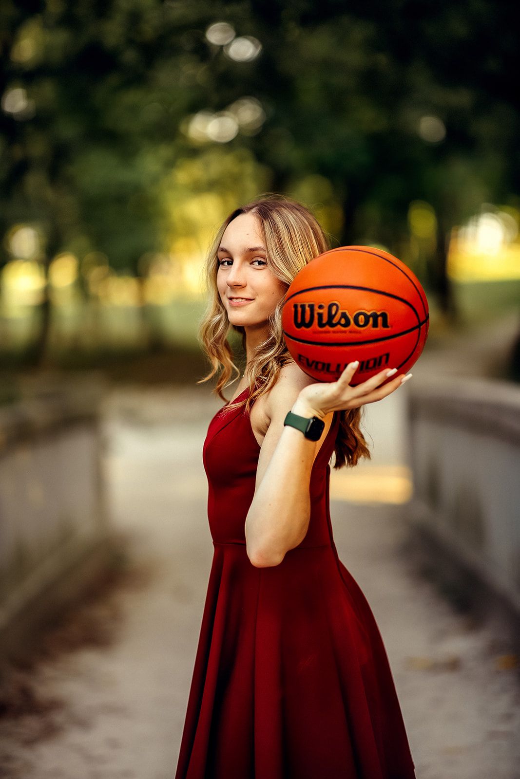 Young woman in red dress holds a Wilson basketball up while posing on a bridge outdoors.
