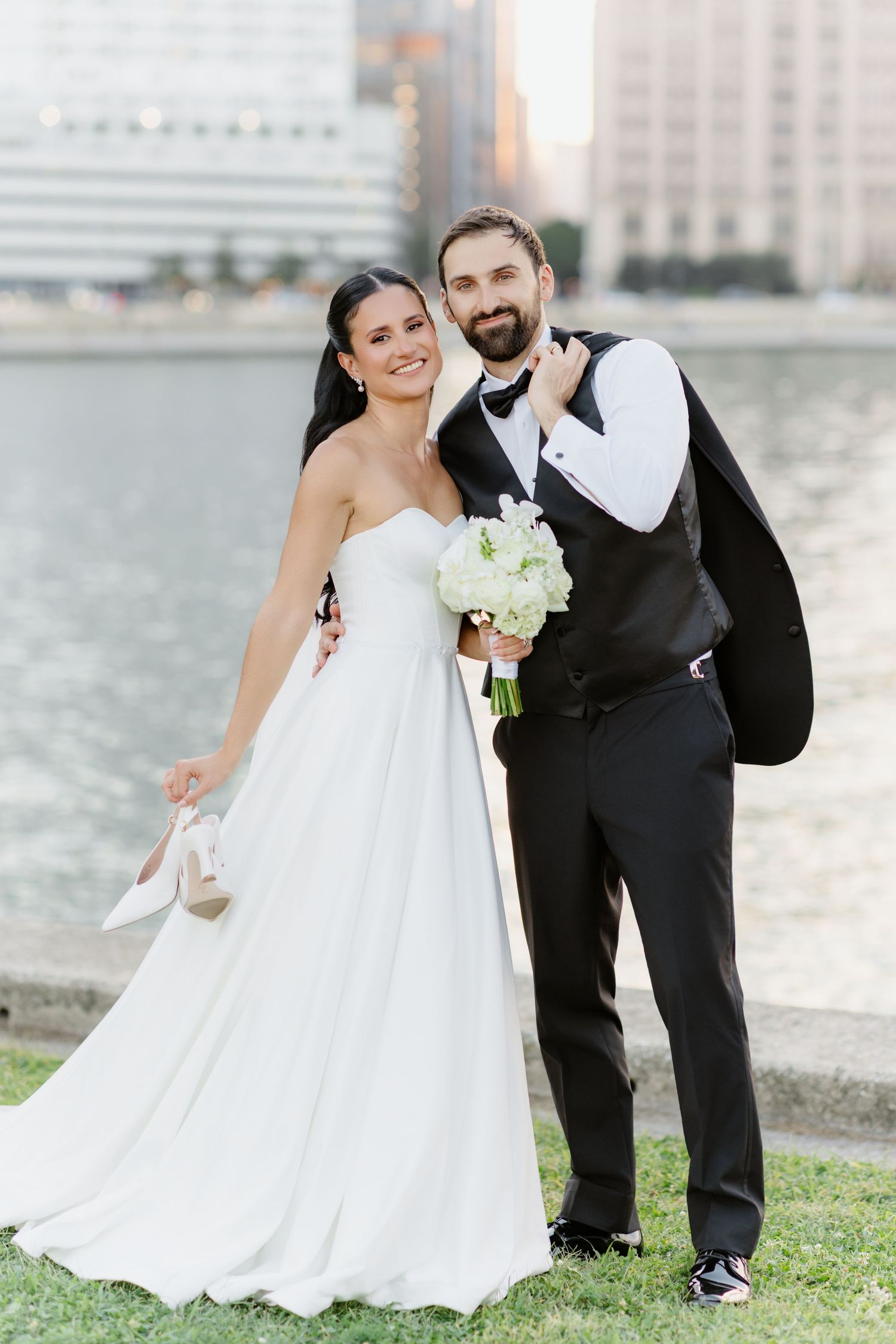 Laughing bride raises tuxedo jacket as groom holds her heels, celebrating by a golden hour city waterfront.
