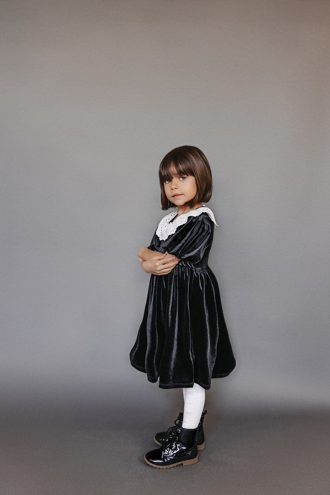Young girl in dark velvet dress with white lace collar, white tights, and black boots, arms crossed, gray background.