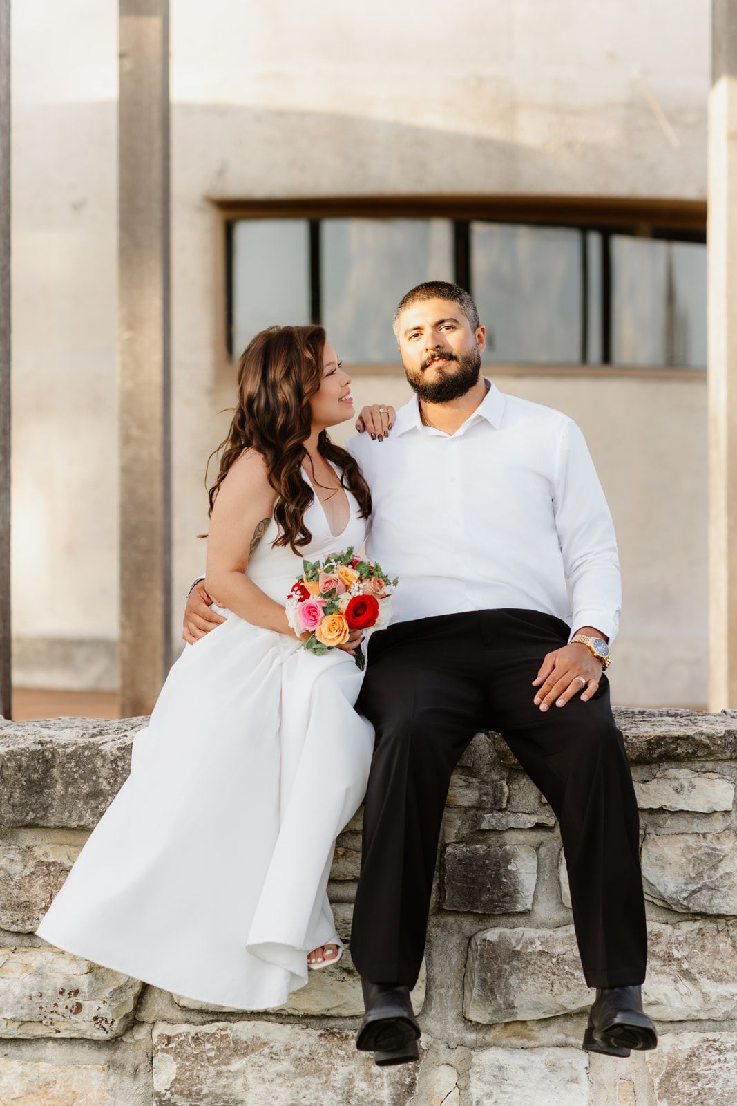 Bride and bearded groom sitting on stone wall with colorful bouquet near building.
