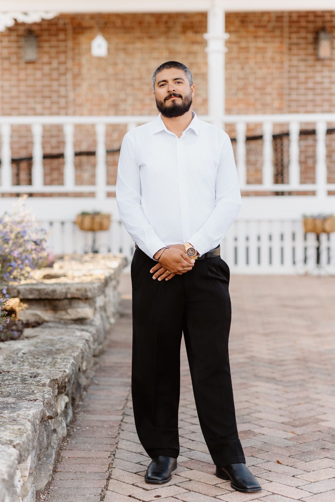 Bearded groom in white shirt and black pants stands with clasped hands outside a brick venue with white railings.