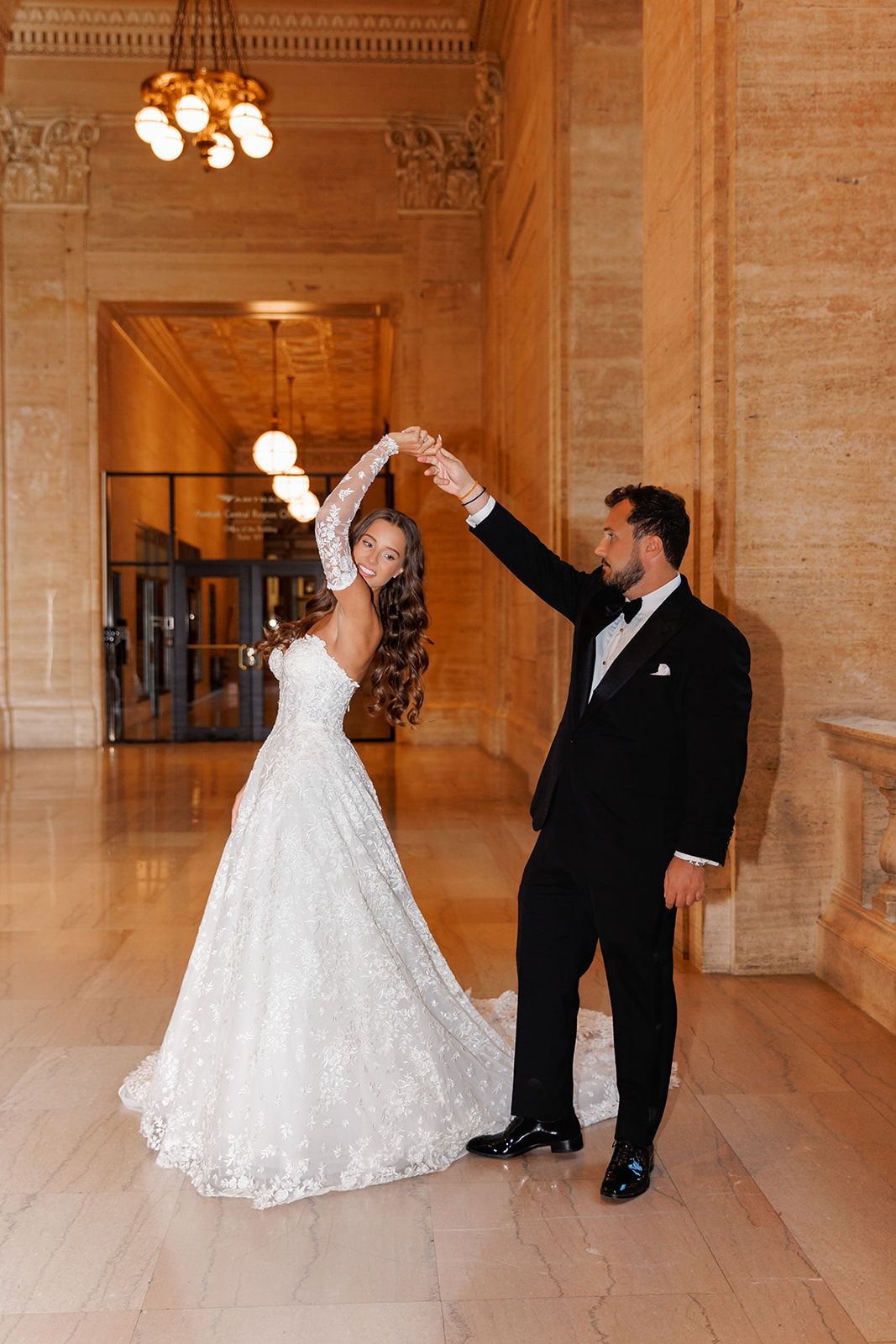 Bride in lace gown twirls as groom in tuxedo spins her in an elegant marble hall with warm chandelier lighting.
