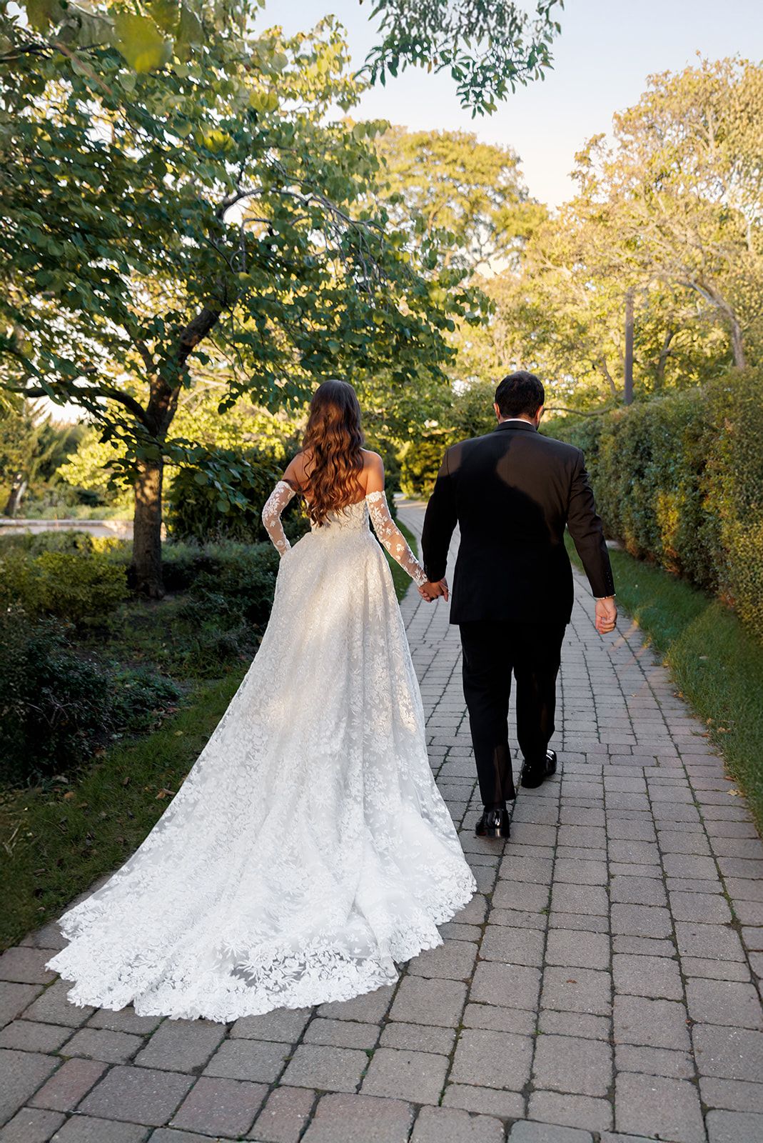 Bride and groom walk hand-in-hand down a tree-lined brick path, showcasing the stunning lace cathedral train of her gown.