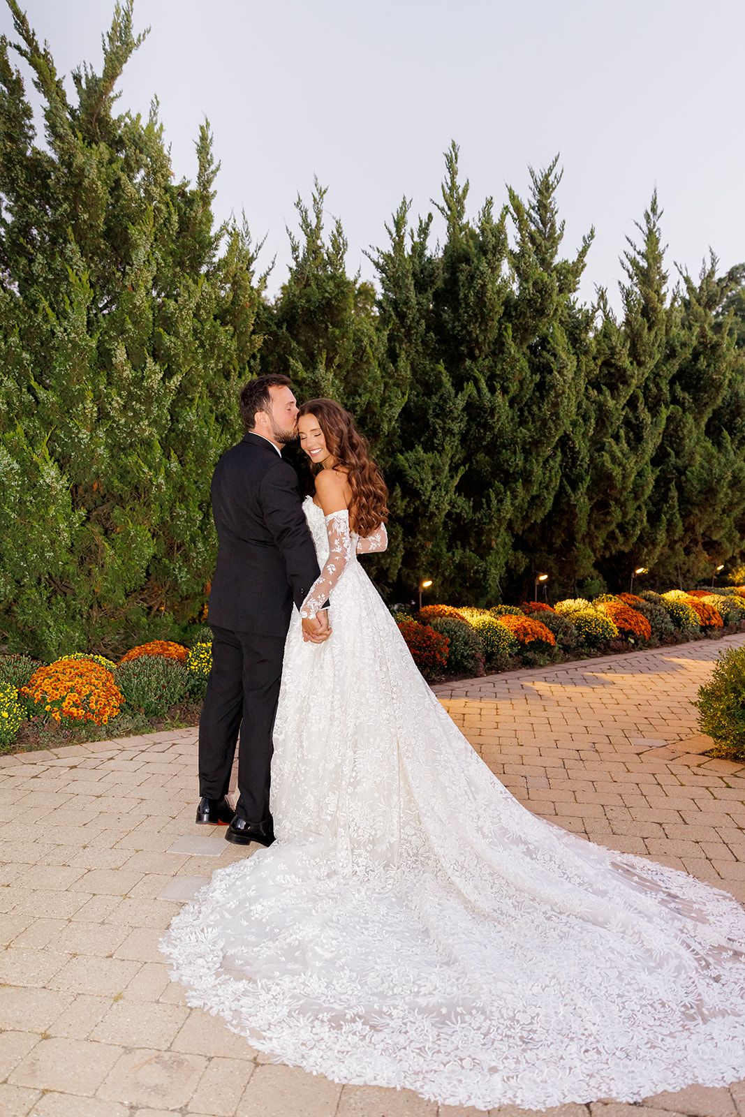 Bride in lace gown with long train kisses groom in black tuxedo outdoors among tall evergreens and colorful mums.