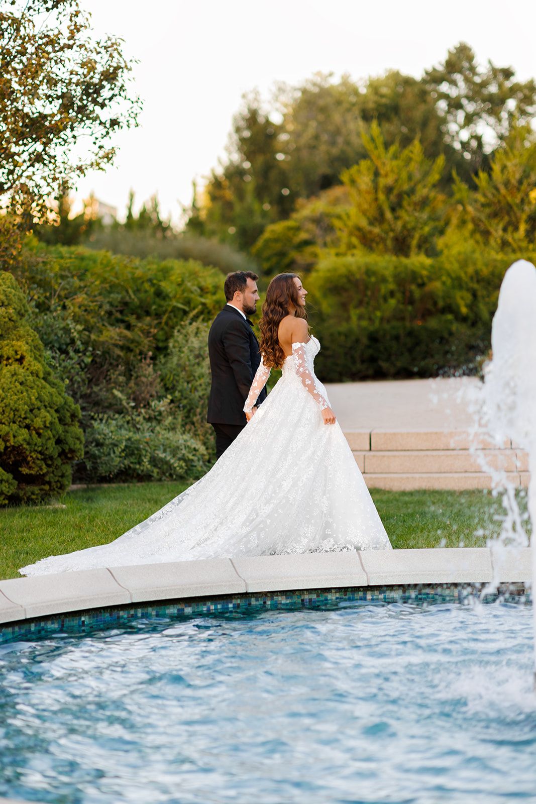 Bride and groom stand by fountain in lush garden, bride wearing stunning lace ball gown with long train.