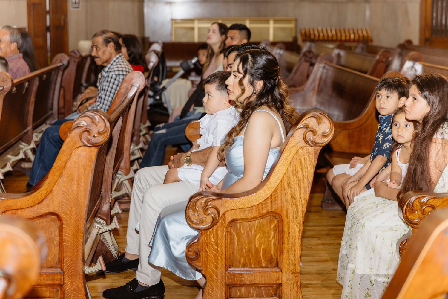 Guests seated in wooden church pews during a wedding ceremony, with the bride visible in a white dress.