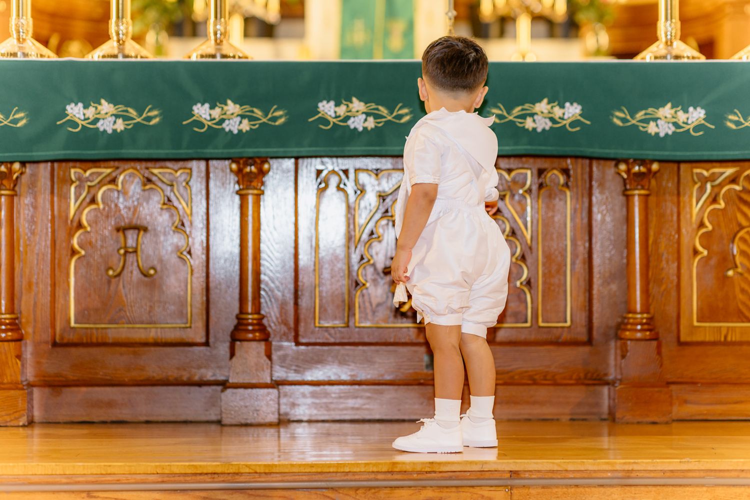 Young child in white outfit standing at ornate wooden church altar with green decorative cloth.