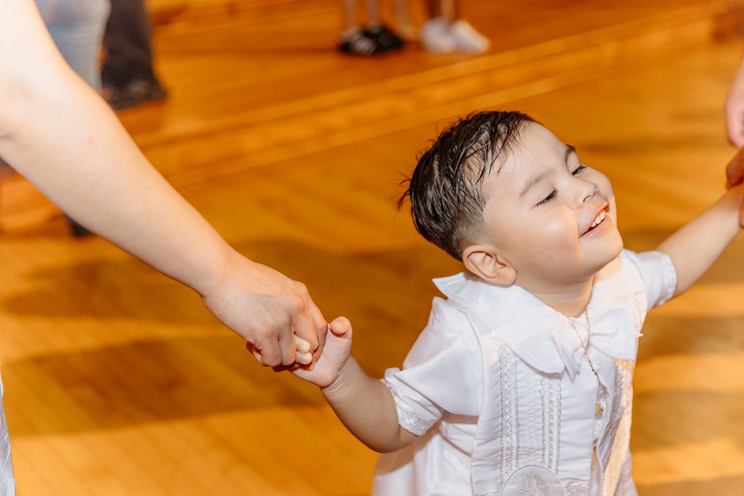 Happy toddler in white outfit holding adult hand, smiling joyfully on wooden dance floor.