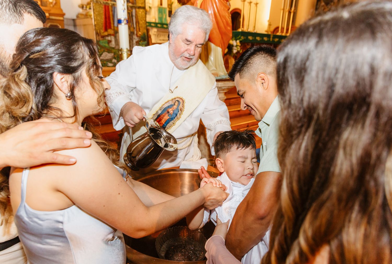 Priest pouring holy water during infant baptism with parents and godparents in decorated Catholic church.