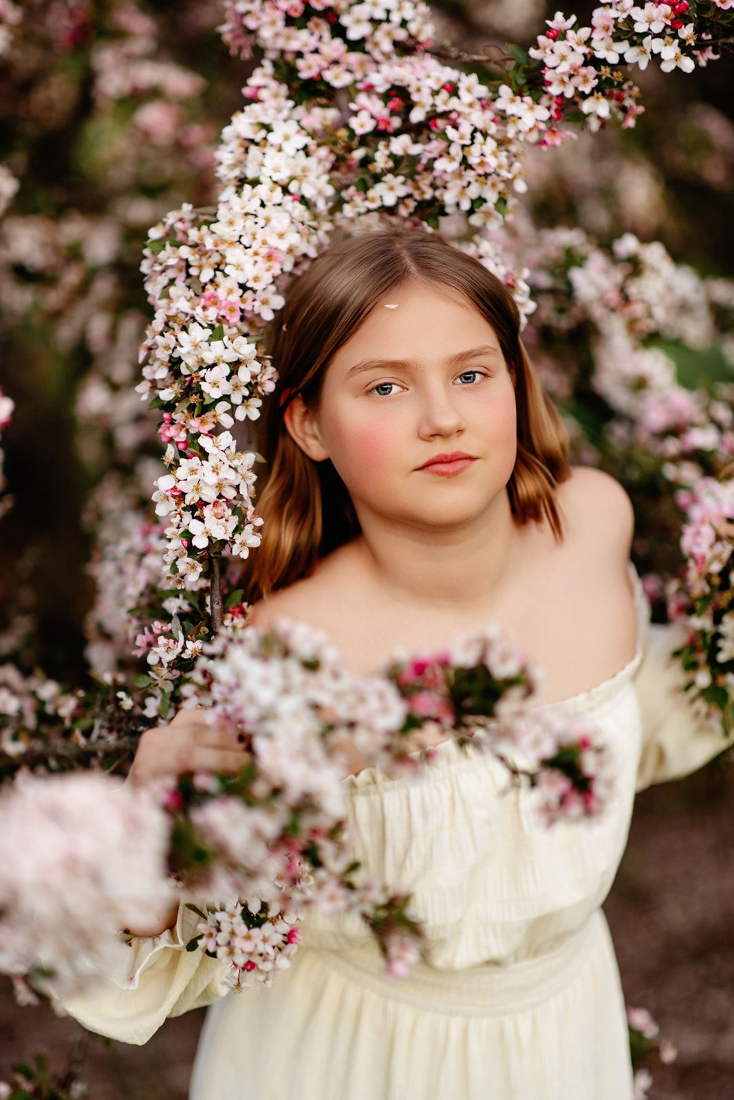 Young girl in off-shoulder cream dress surrounded by pink and white spring blossoms on a flowering tree.